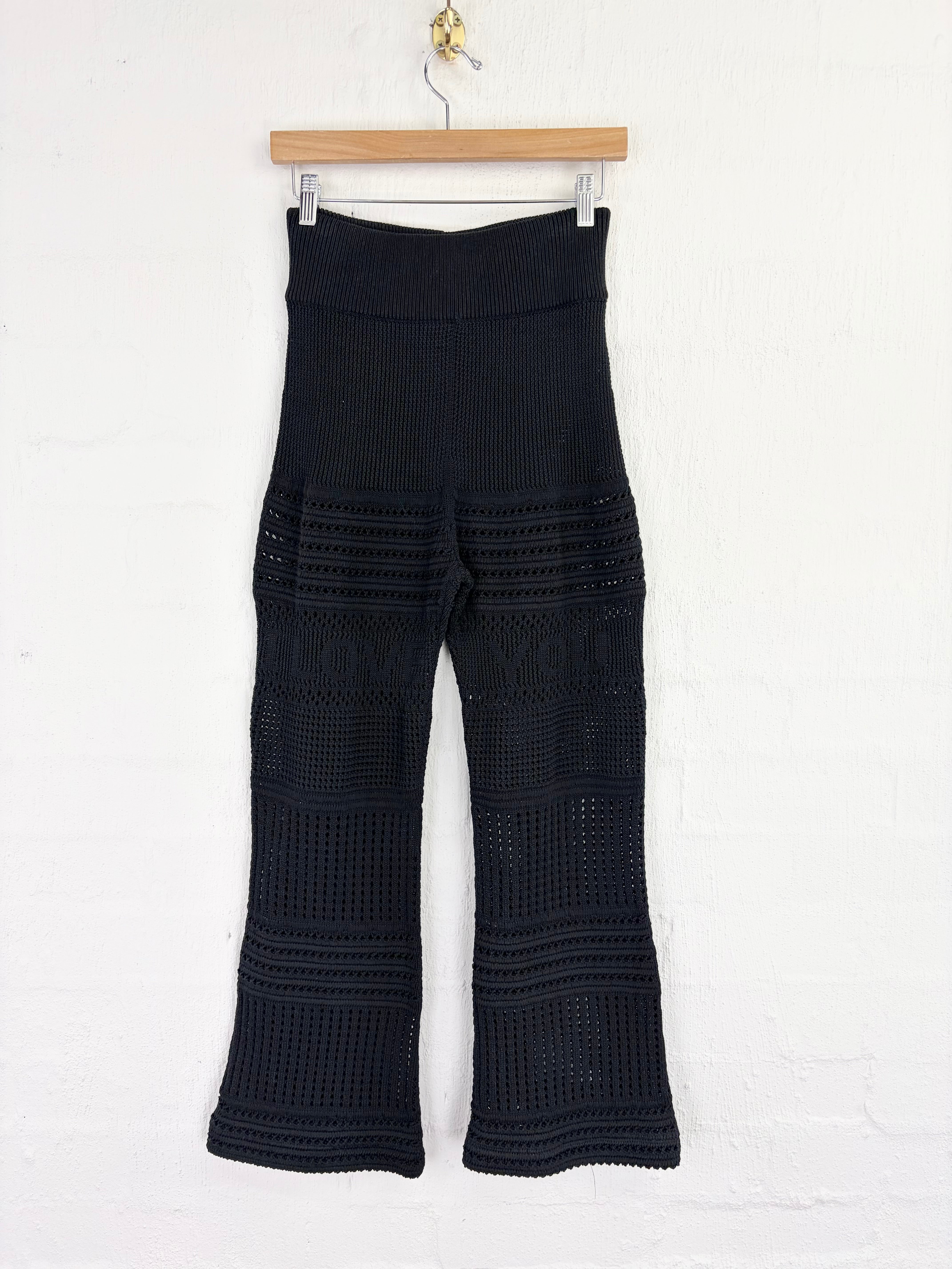 Black crochet knit flare pants hanging from a wooden hanger against a white brick wall.
