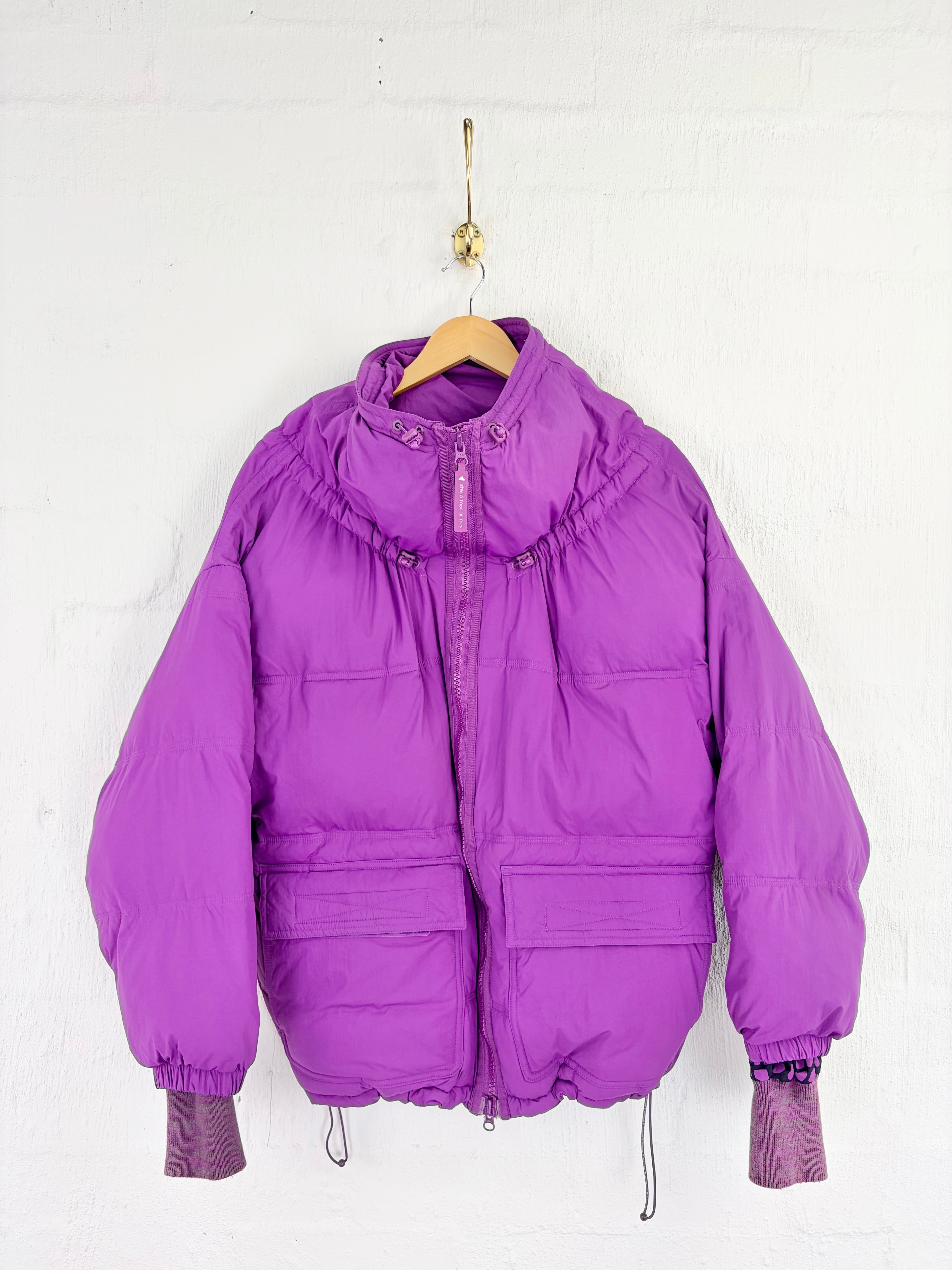 A bright purple puffer jacket hanging on a wooden hanger against a white brick wall.