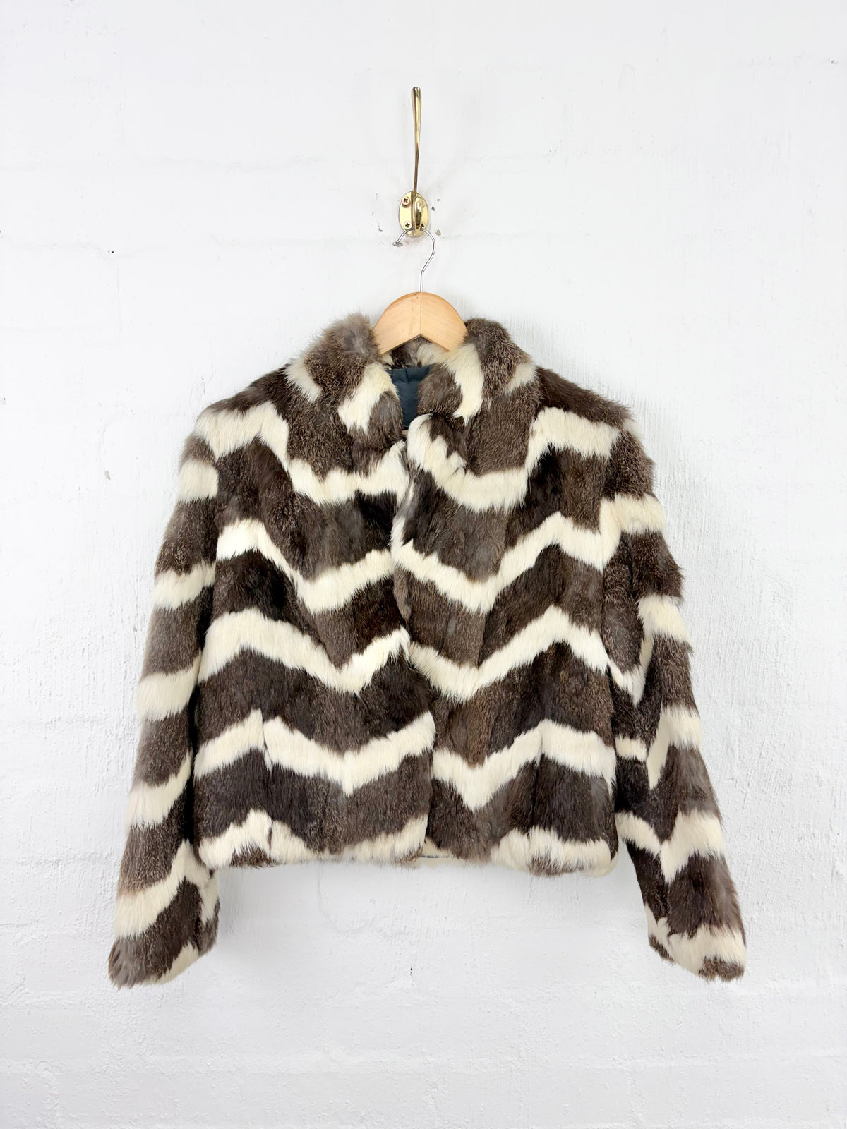 A cropped fur jacket with a brown and cream chevron pattern, hanging on a wooden hanger against a white painted brick wall.
