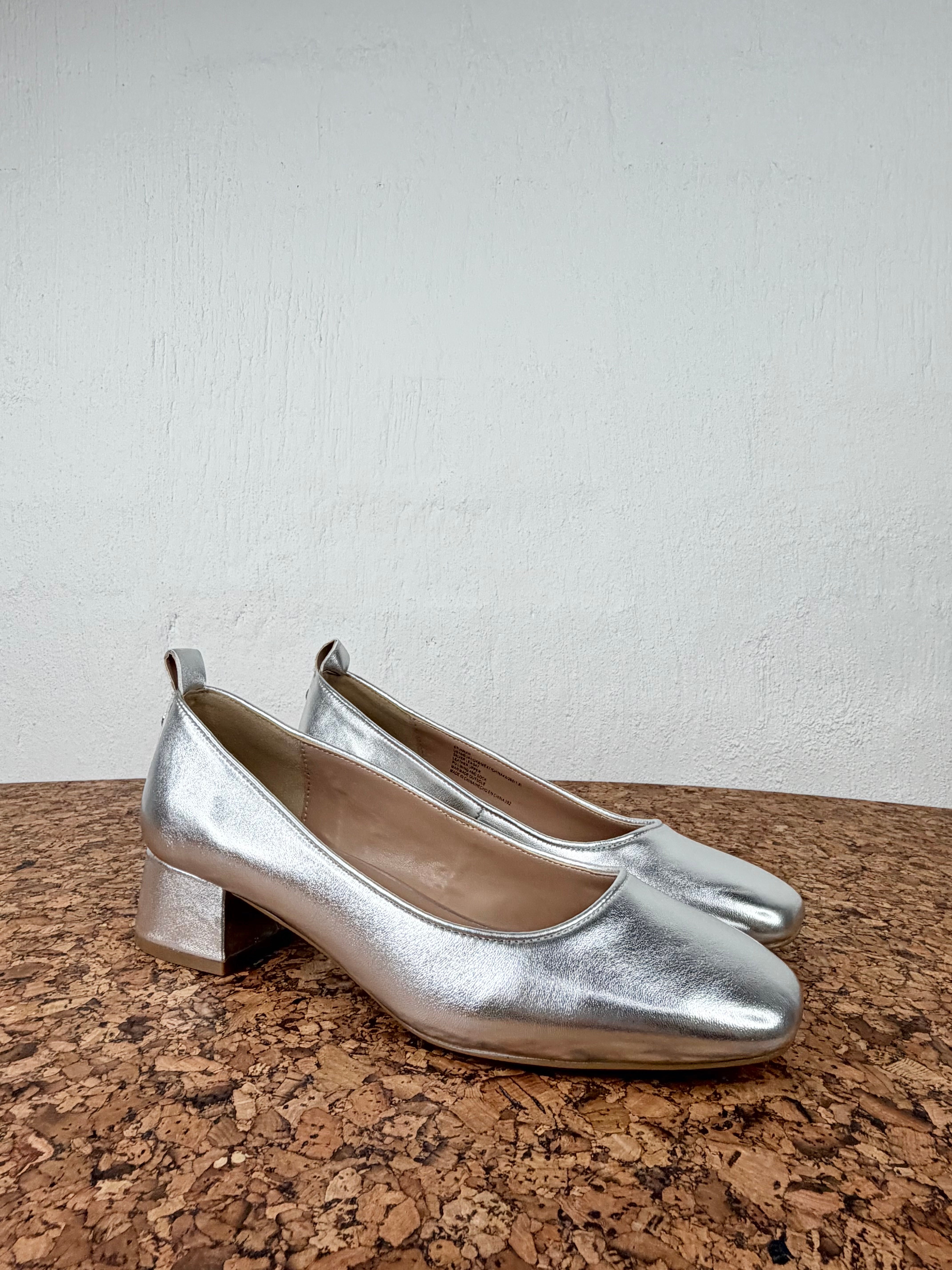 A pair of metallic silver leather pump shoes with low block heels, displayed on a brown cork surface against a light gray textured wall.