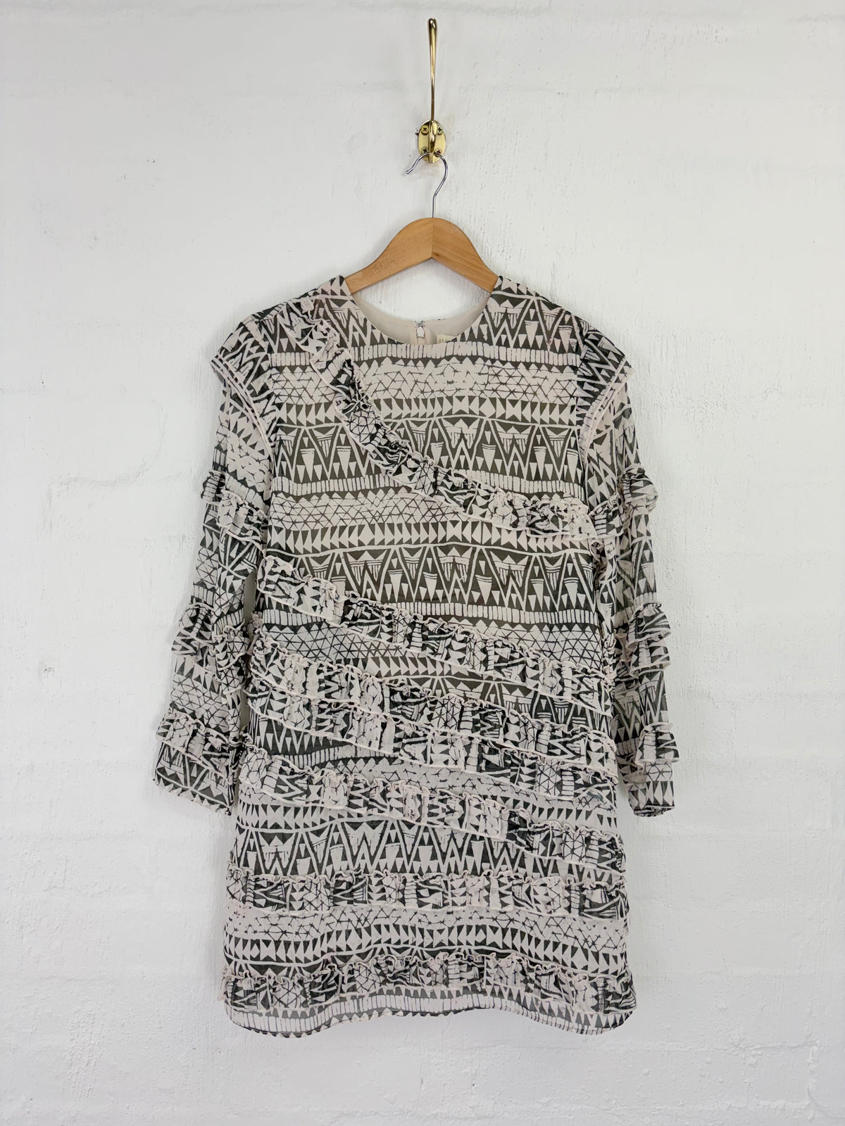 A black and white patterned knit tunic top with tiered ruffle sleeves, hanging on a wooden hanger against a white painted brick wall.