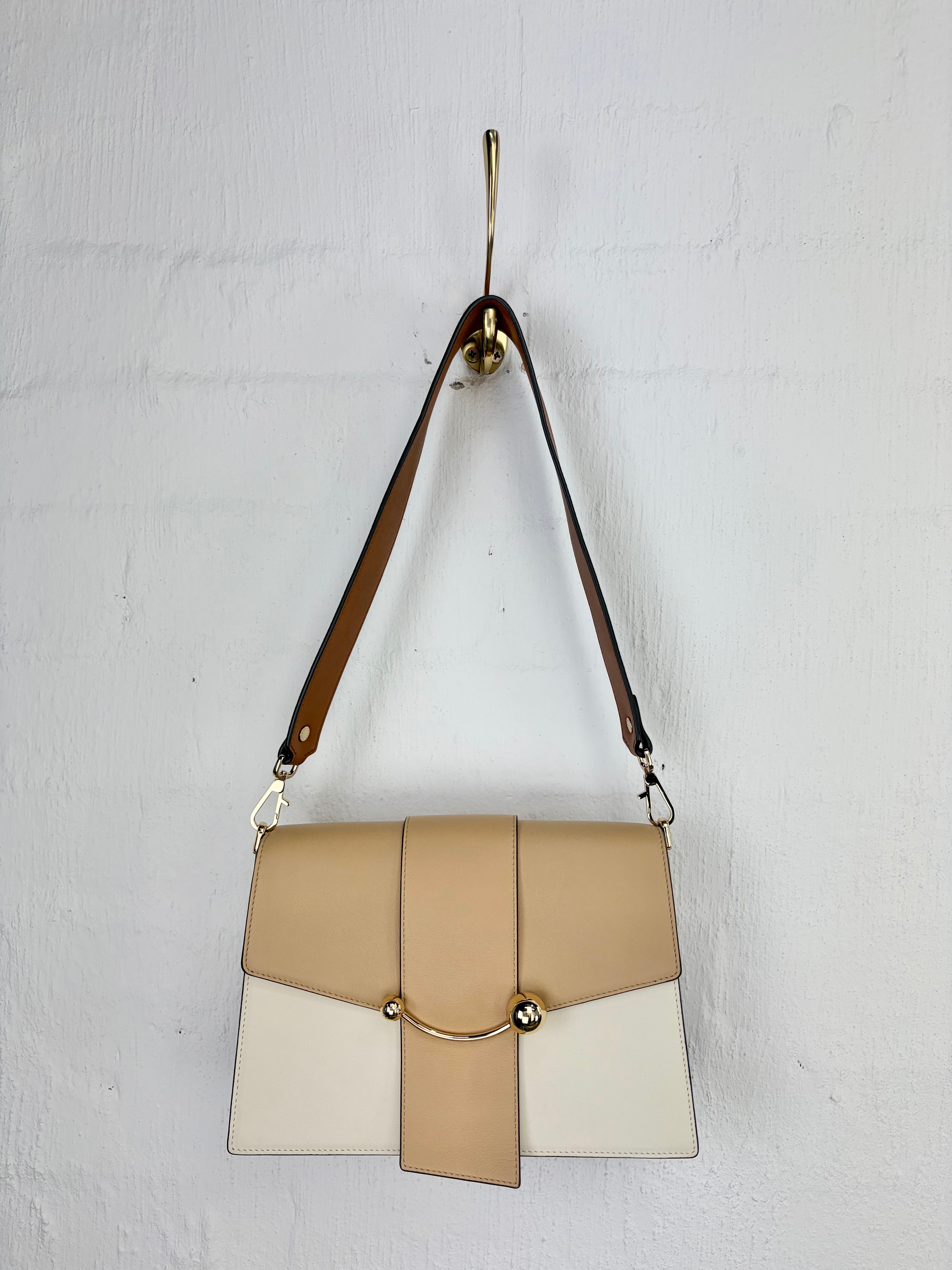 A structured leather shoulder bag in beige, cream, and white with a brown strap, hanging from a gold hook against a white painted brick wall.