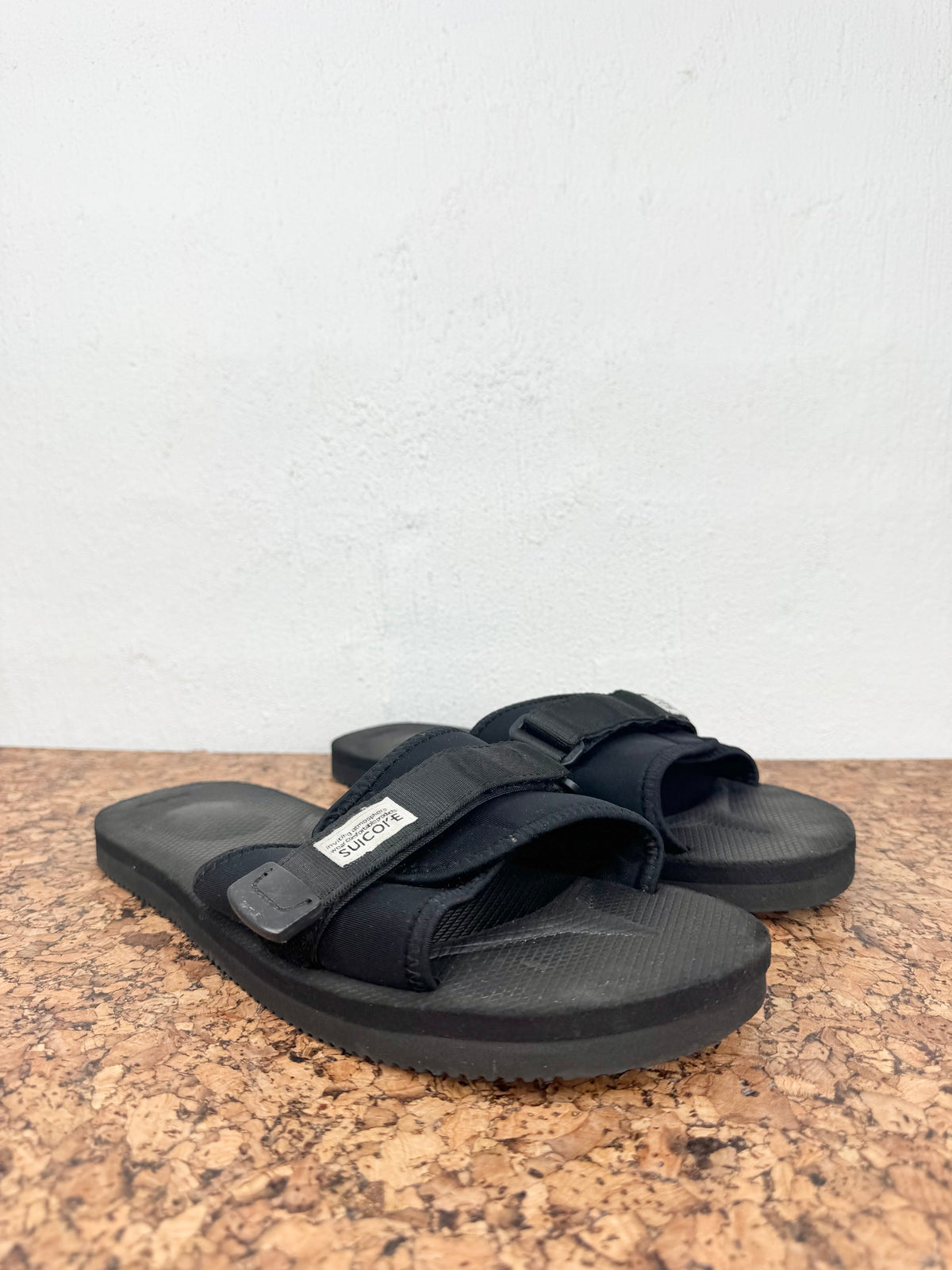 A pair of black slide sandals with adjustable hook-and-loop straps and a textured sole, resting on a brown speckled surface against a white wall.