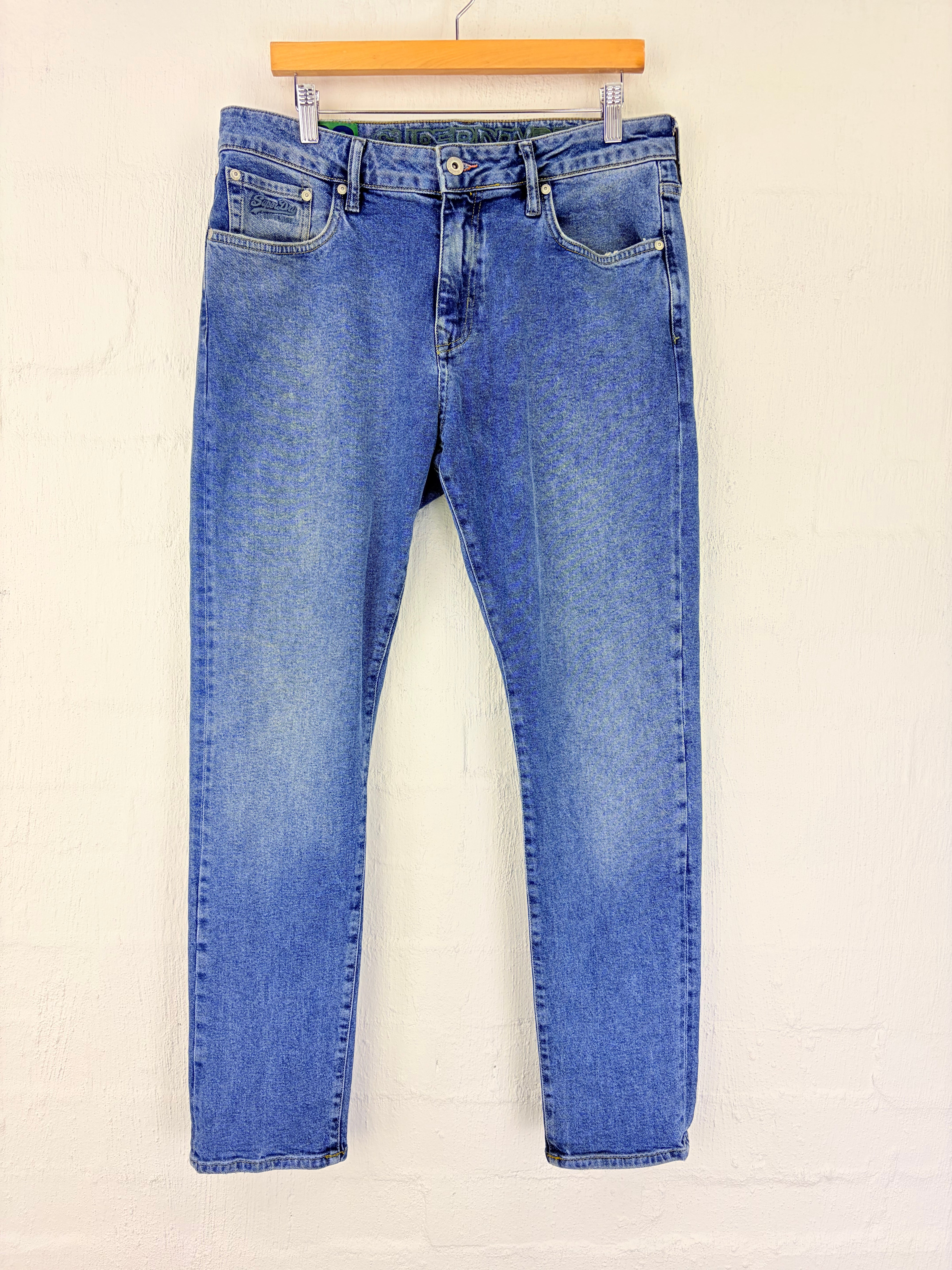 A pair of medium wash blue denim jeans hanging from a wooden hanger against a white painted brick wall.