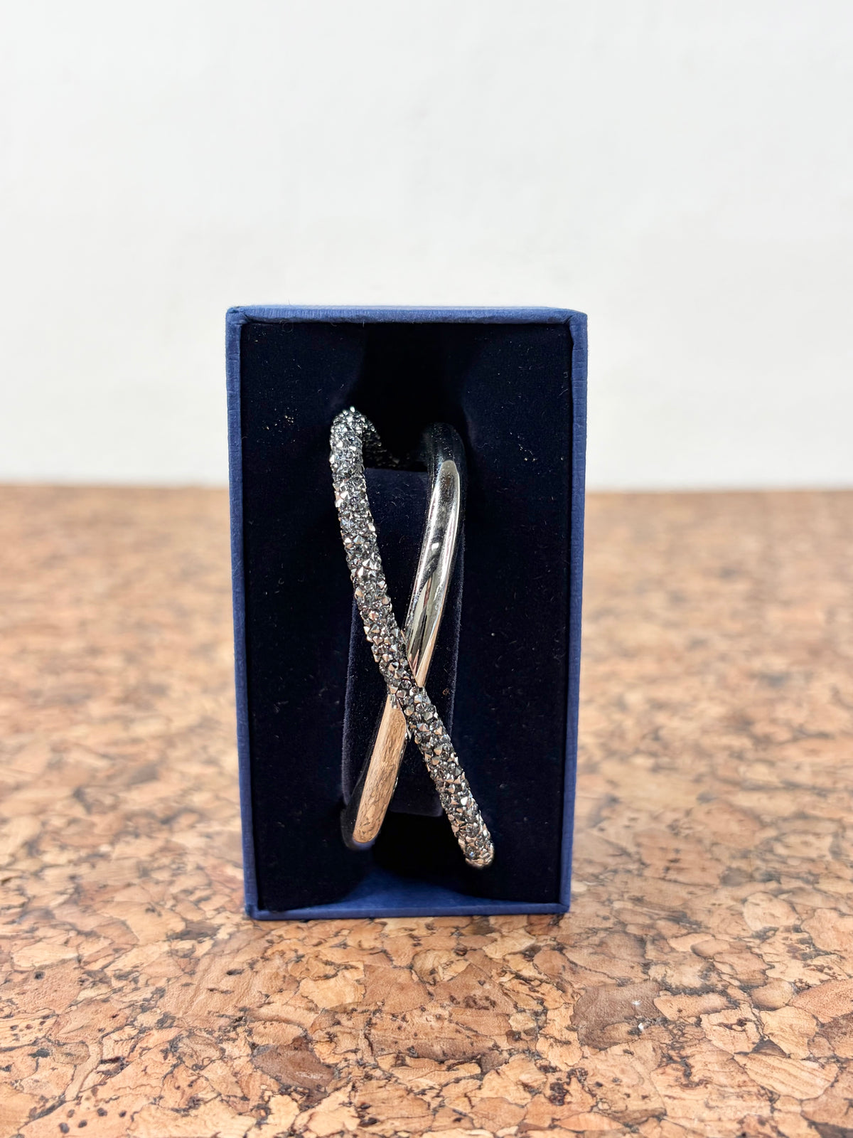 A silver-toned double-band ring with one band paved with small clear stones and the other plain, displayed inside an open dark blue jewelry box on a cork surface against a white wall.