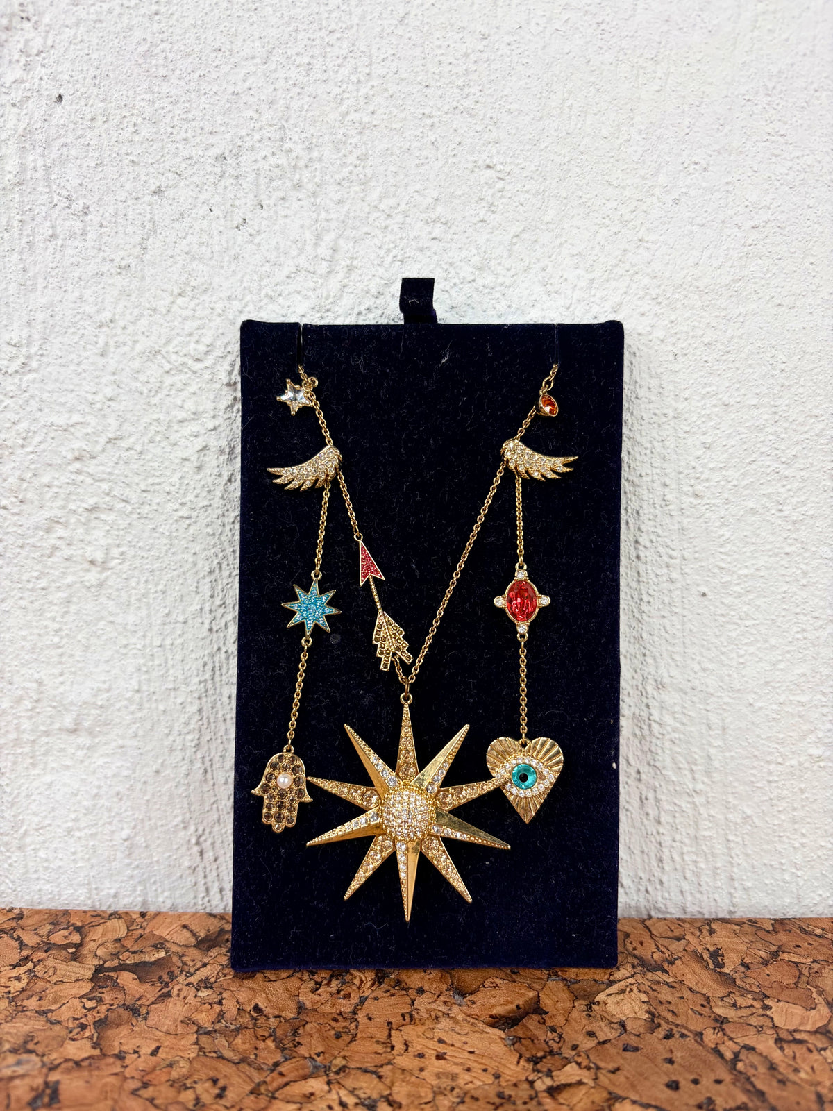 A gold-toned multi-charm necklace displayed on a black velvet jewelry card, featuring a large starburst pendant, angel wings, a hamsa hand, a heart with a turquoise stone, and other small decorative charms, set against a white textured wall and cork surface.