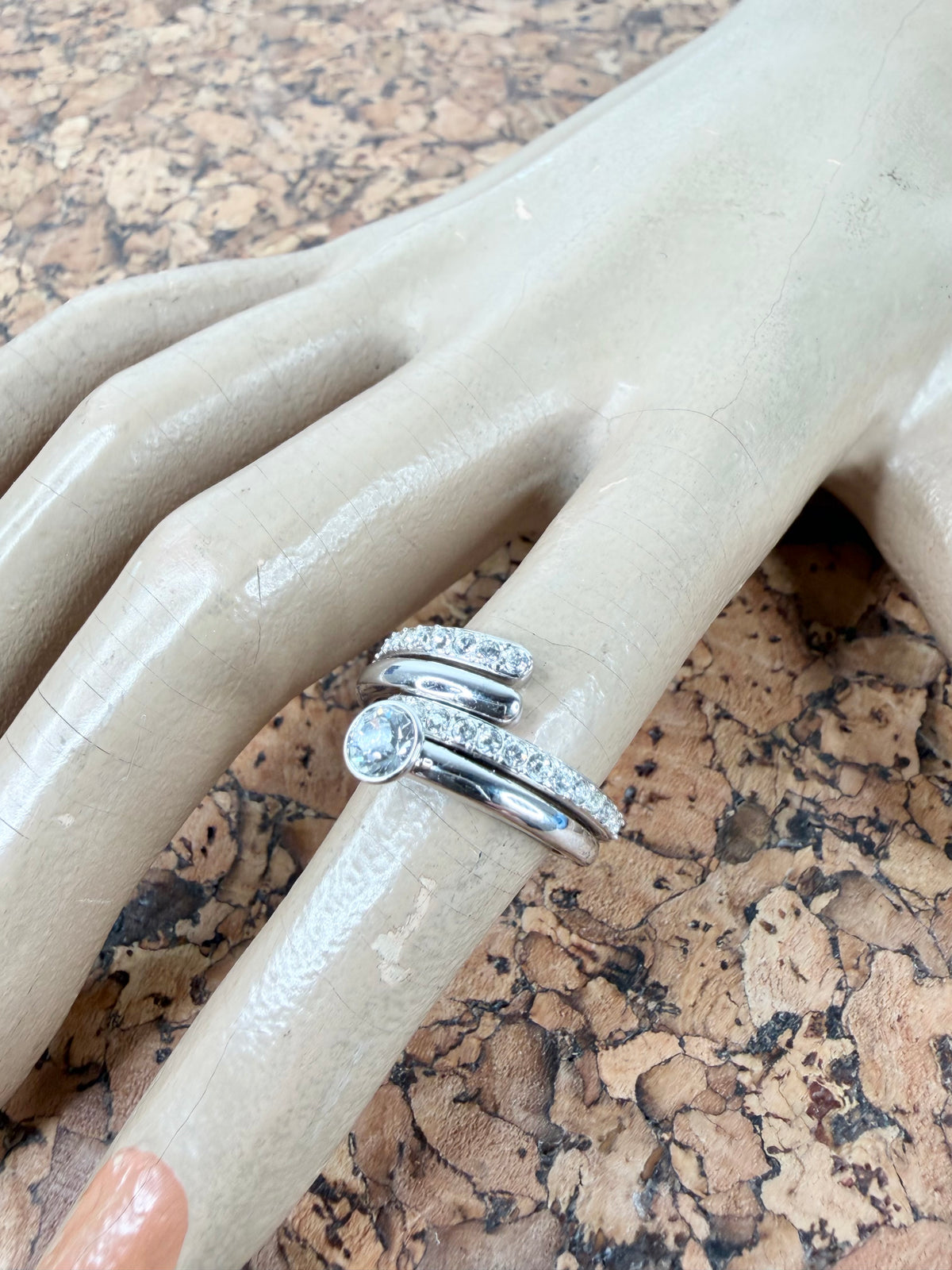 A silver-toned three-band ring set displayed on a beige mannequin hand over a cork surface. The set includes a band with a round-cut clear stone and pavé accents, a textured band, and a slim band.