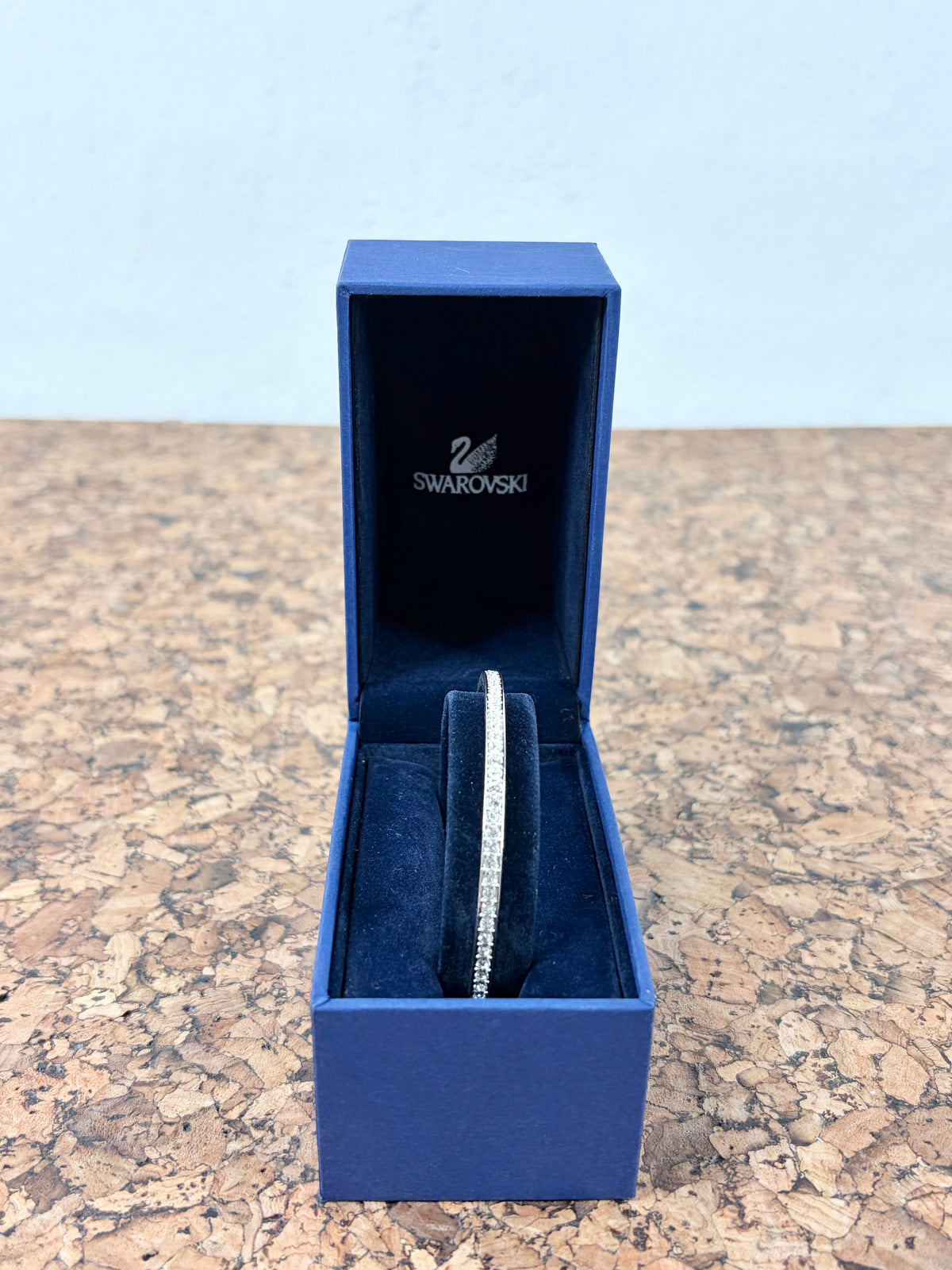 A Swarovski-branded blue jewelry box opened to display a slim silver-toned bracelet with clear stones, set on a speckled countertop against a light blue wall.