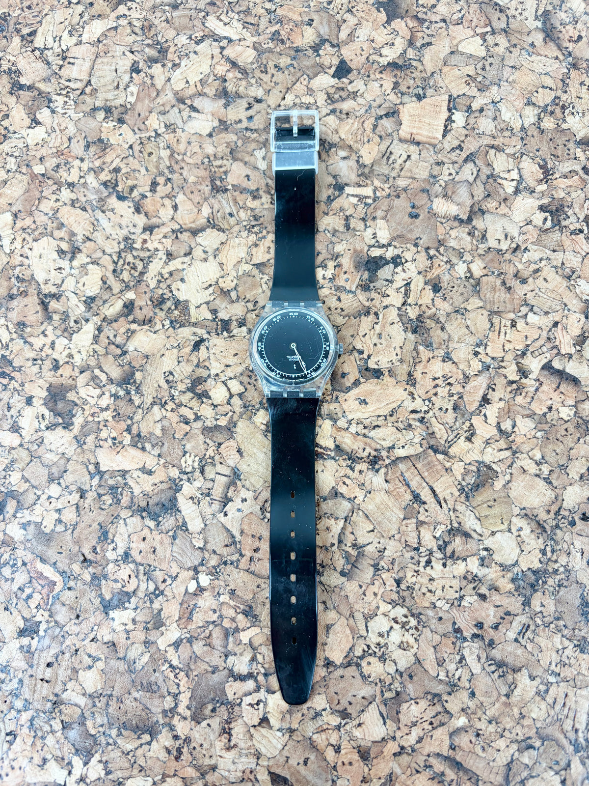 A Swatch wristwatch with a black rubber strap and a silver-tone case, laid flat on a textured cork surface.