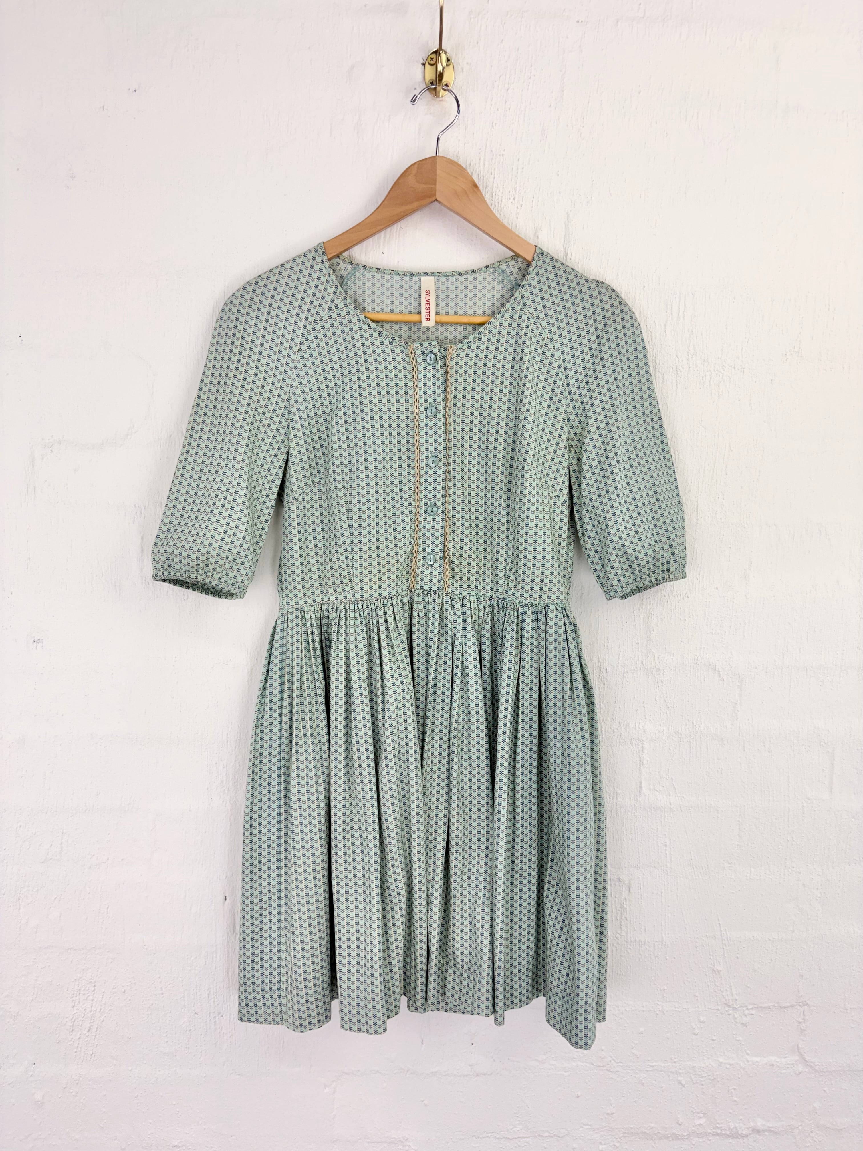 A sage green and white patterned short-sleeve dress hanging on a wooden hanger against a white brick wall.