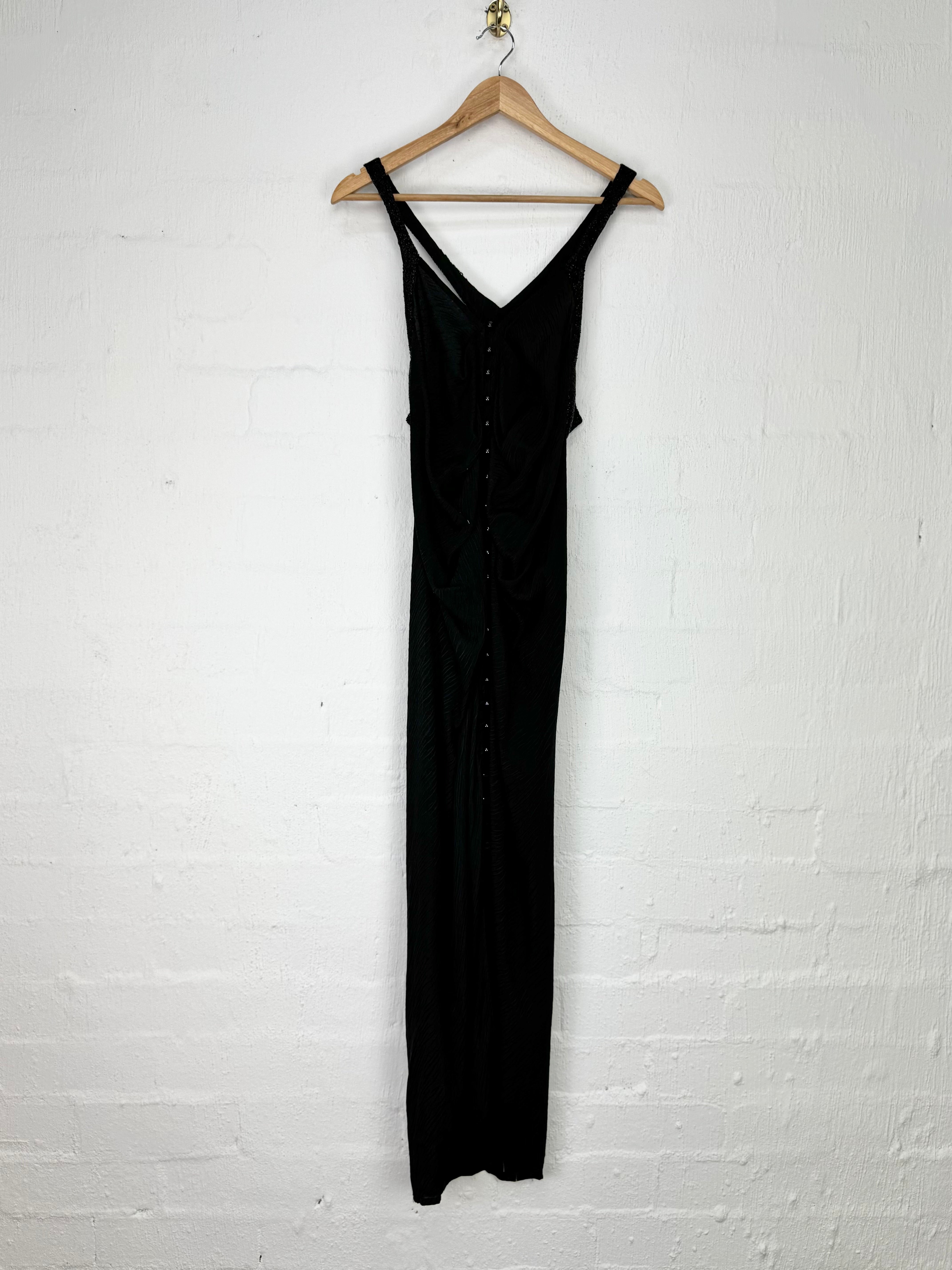 A black sleeveless maxi dress with a V-neckline and button-front detail, hanging on a wooden hanger against a white painted brick wall.
