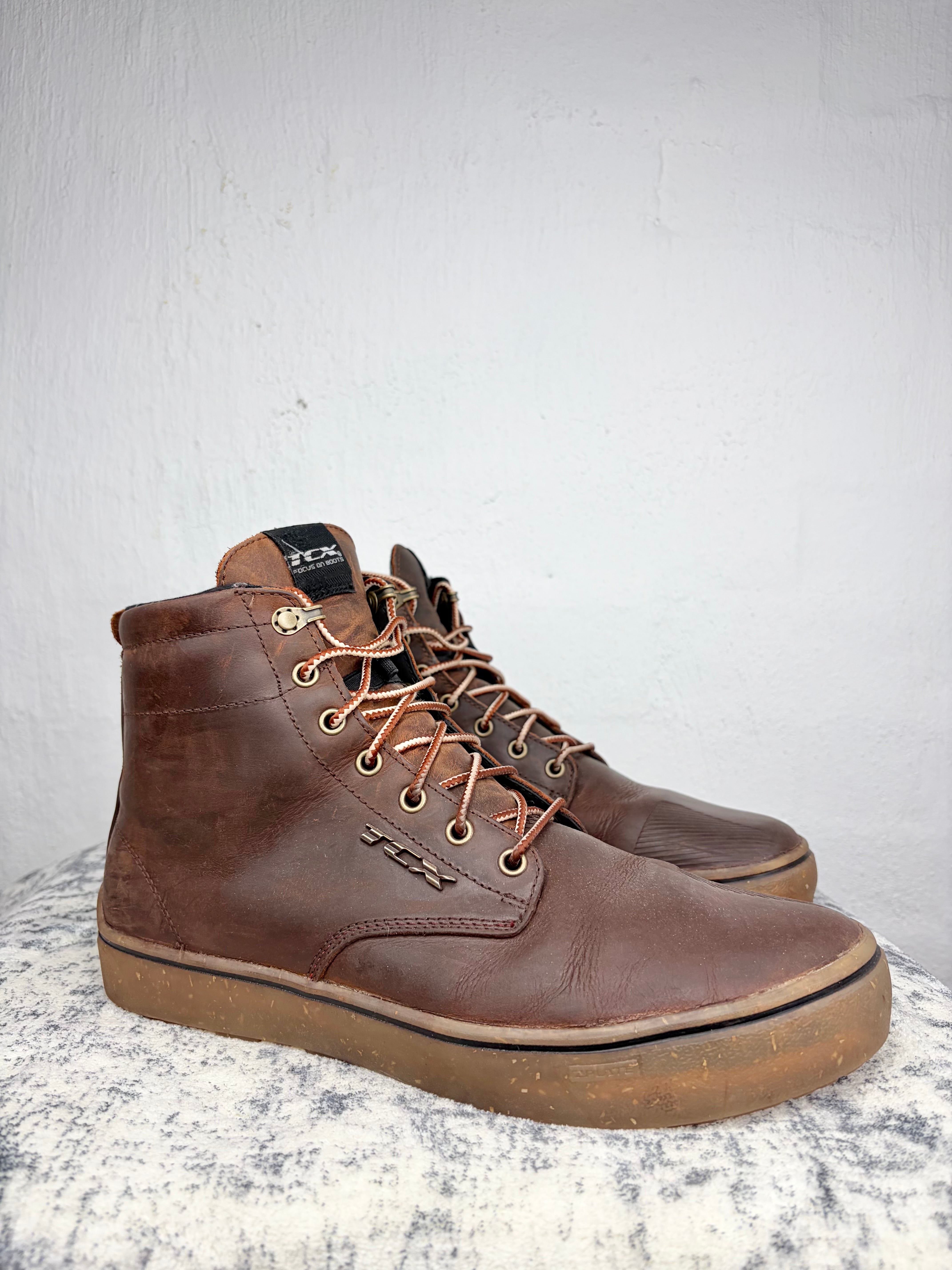 A pair of brown leather high-top boots with tan laces and gum soles, displayed on a textured gray surface against a white wall.