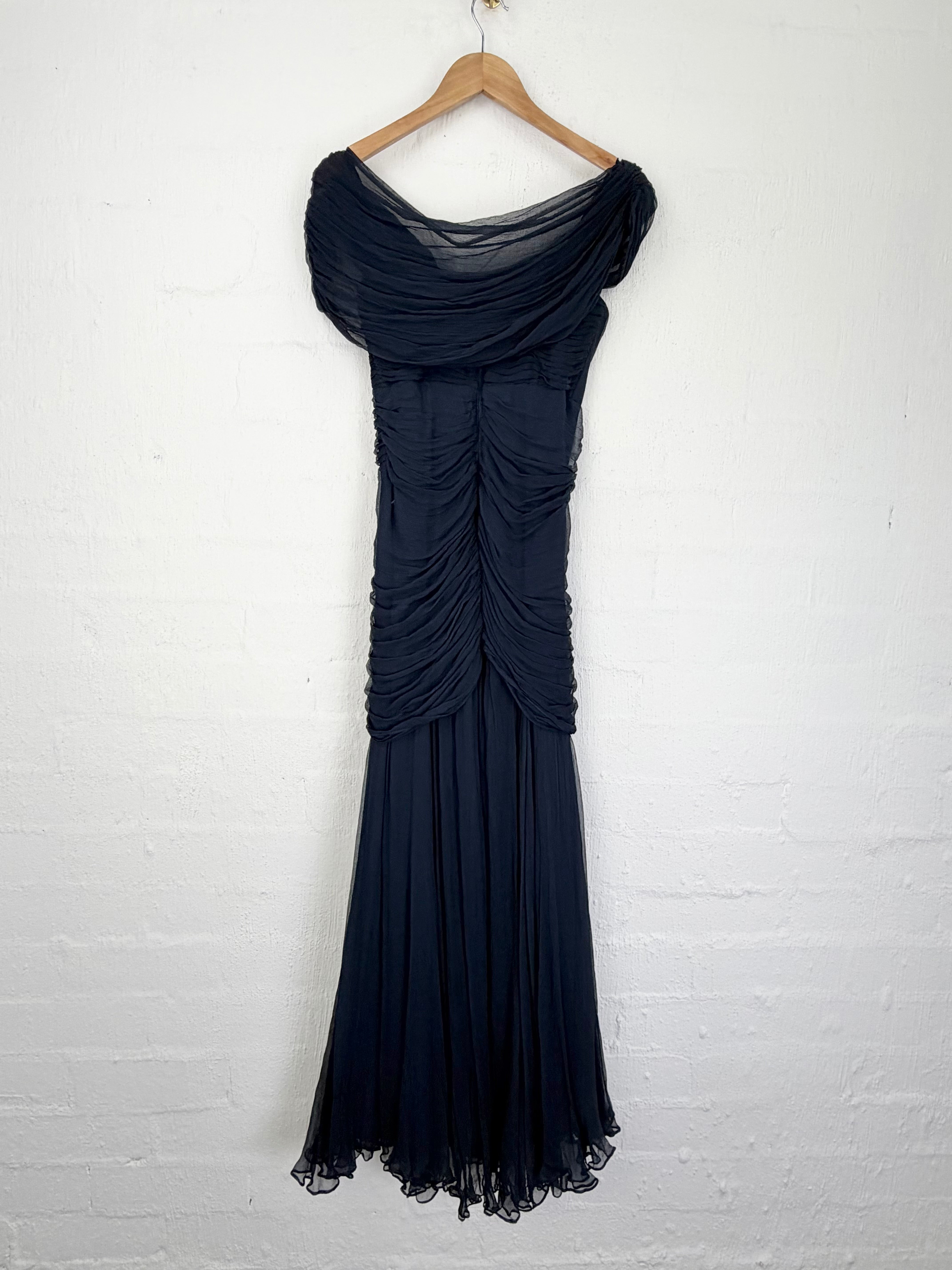 A dark navy blue off-the-shoulder gown with dramatic draped ruching, hanging from a wooden hanger against a white painted brick wall.