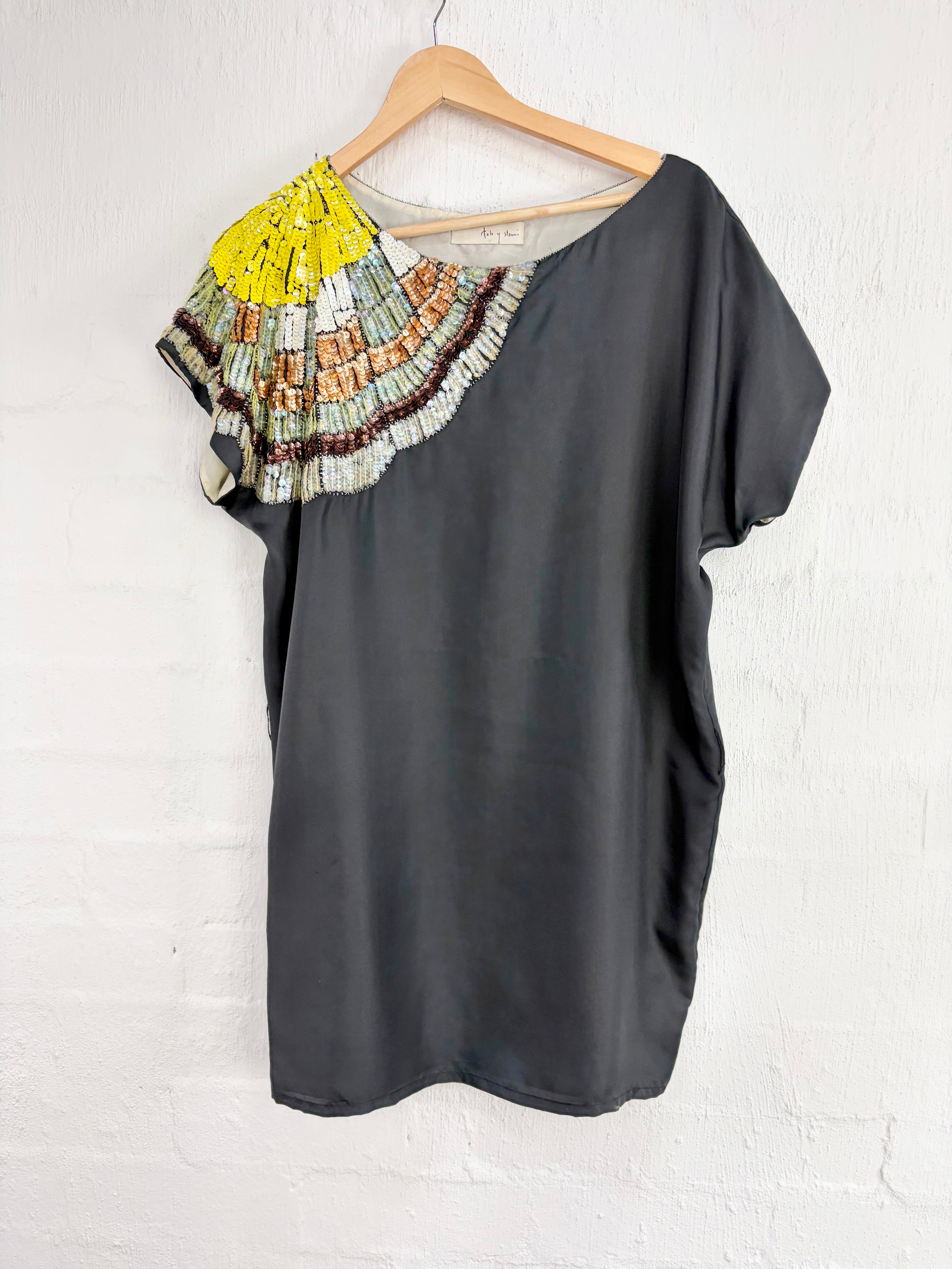 A dark charcoal gray short-sleeve blouse with a colorful pleated yoke detail on the left shoulder, hanging on a wooden hanger against a white painted brick wall.