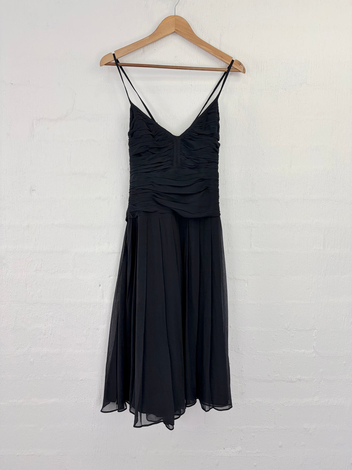 A black sleeveless midi dress with thin spaghetti straps, a ruched sweetheart bodice, and a flowing chiffon skirt, hanging on a wooden hanger against a white painted brick wall.
