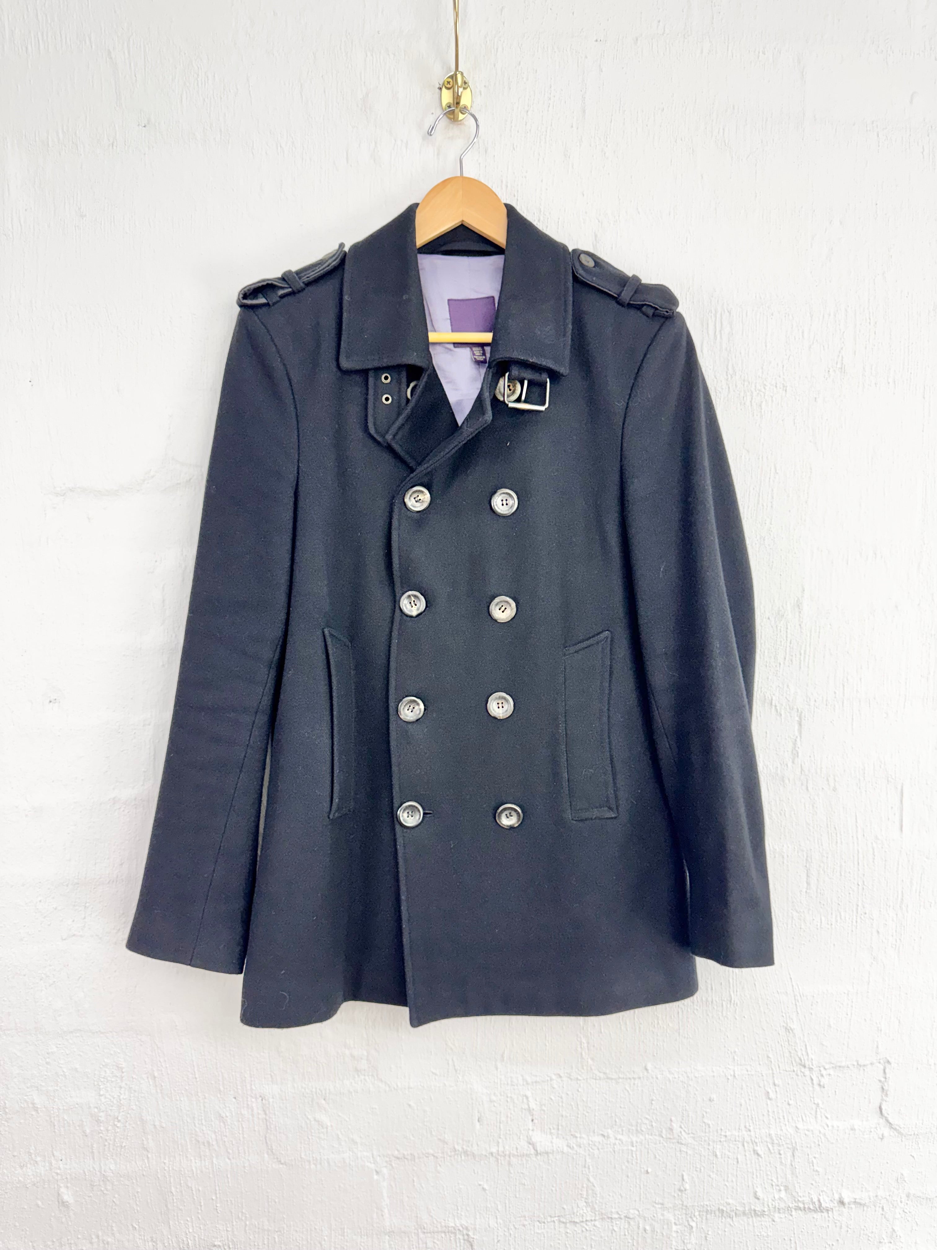 A dark charcoal gray double-breasted military-style coat with silver buttons, hanging on a wooden hanger against a white painted brick wall.
