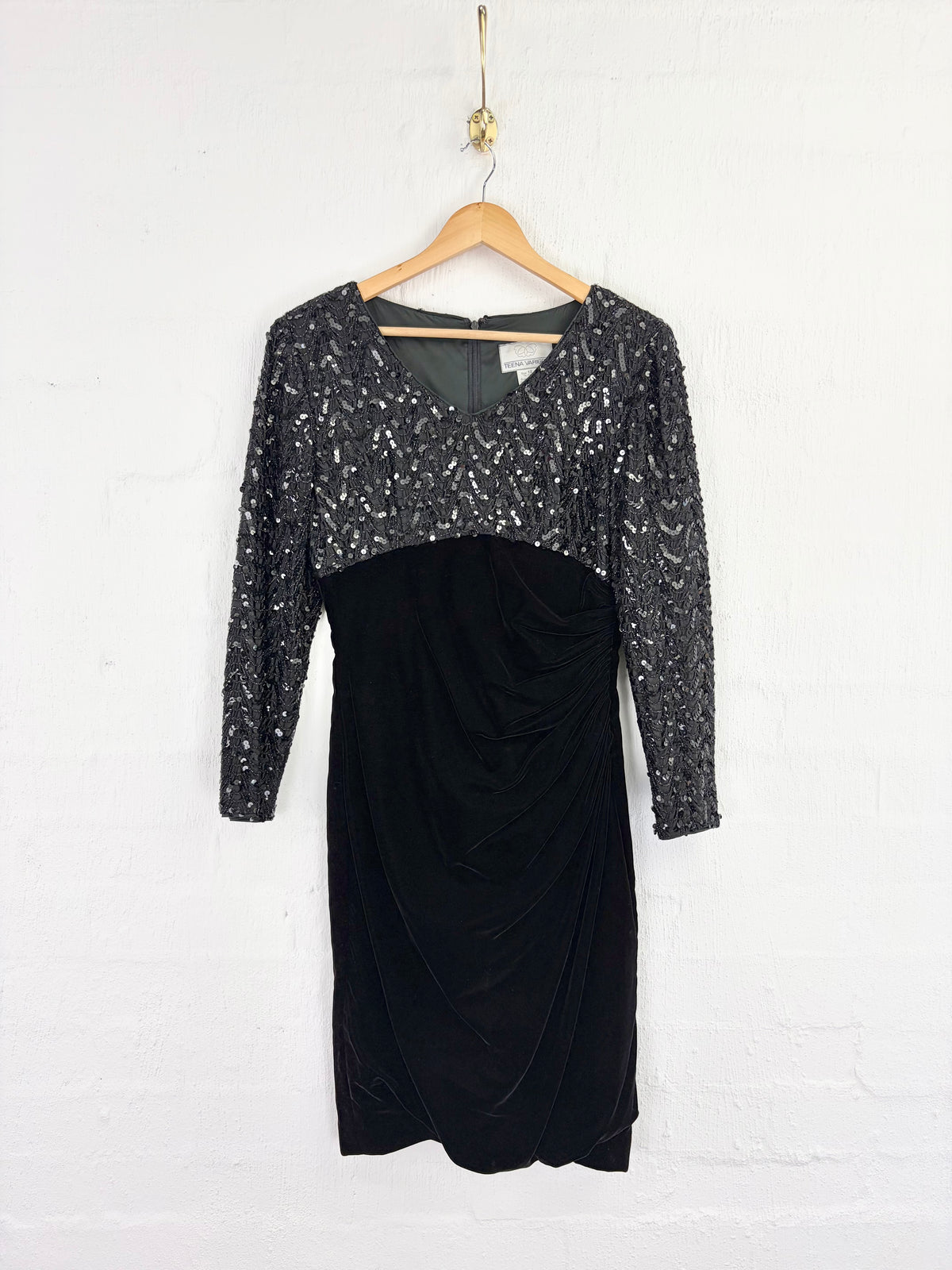 A black long-sleeve dress with a sparkling silver sequined bodice and sleeves, hanging on a wooden hanger against a white brick wall.