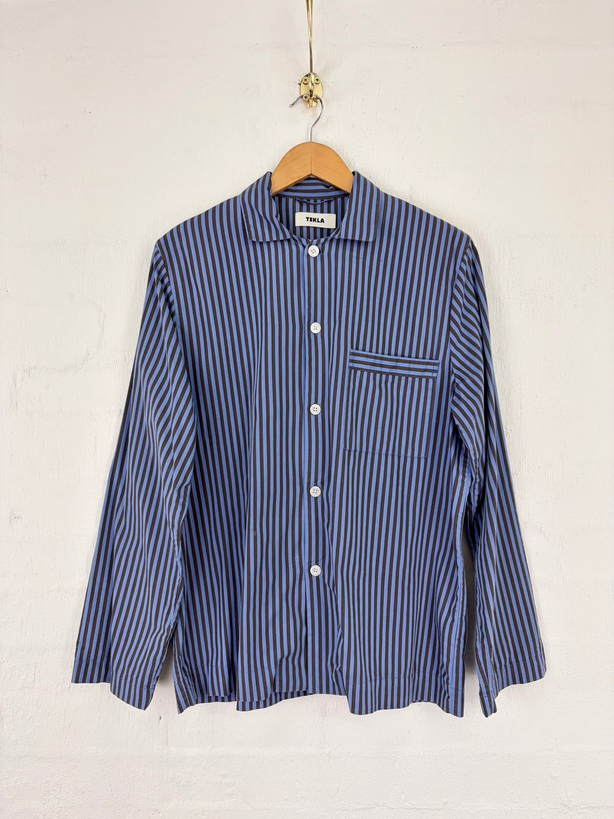 A blue and white vertically striped long-sleeve button-up shirt hanging on a wooden hanger against a white textured wall.