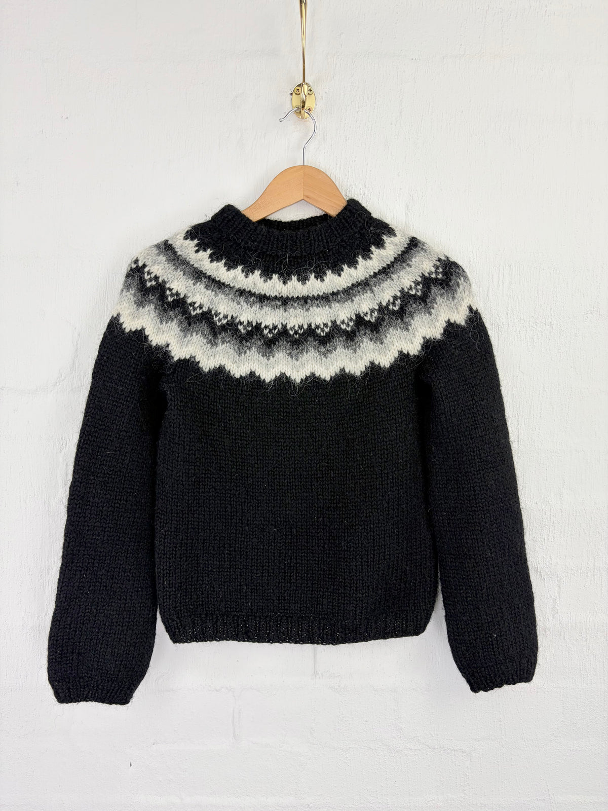 A black knit sweater with a cream and gray Fair Isle yoke pattern, hanging on a wooden hanger against a white brick wall.