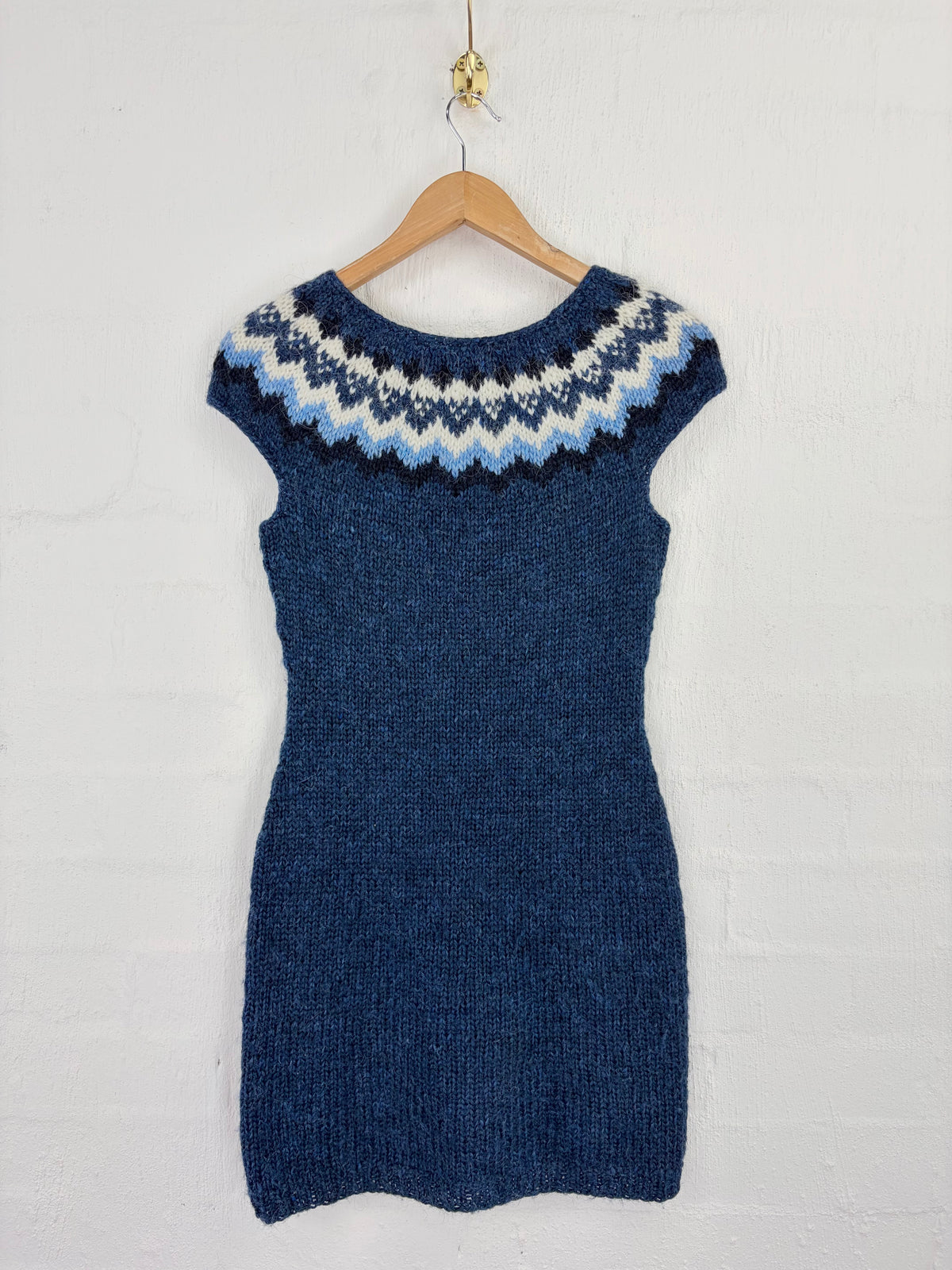 A hand-knit short-sleeve dress in dark blue with a Fair Isle yoke pattern in navy, white, and light blue, hanging on a wooden hanger against a white brick wall.