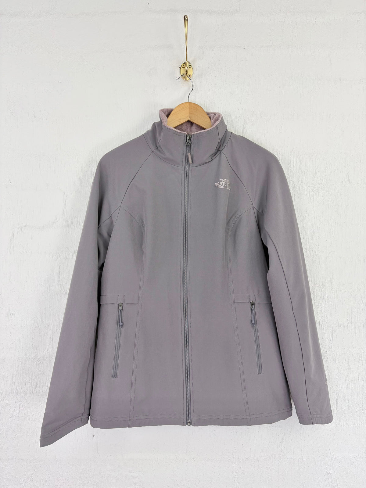 A gray zip-up jacket by The North Face hanging on a wooden hanger against a white textured wall.