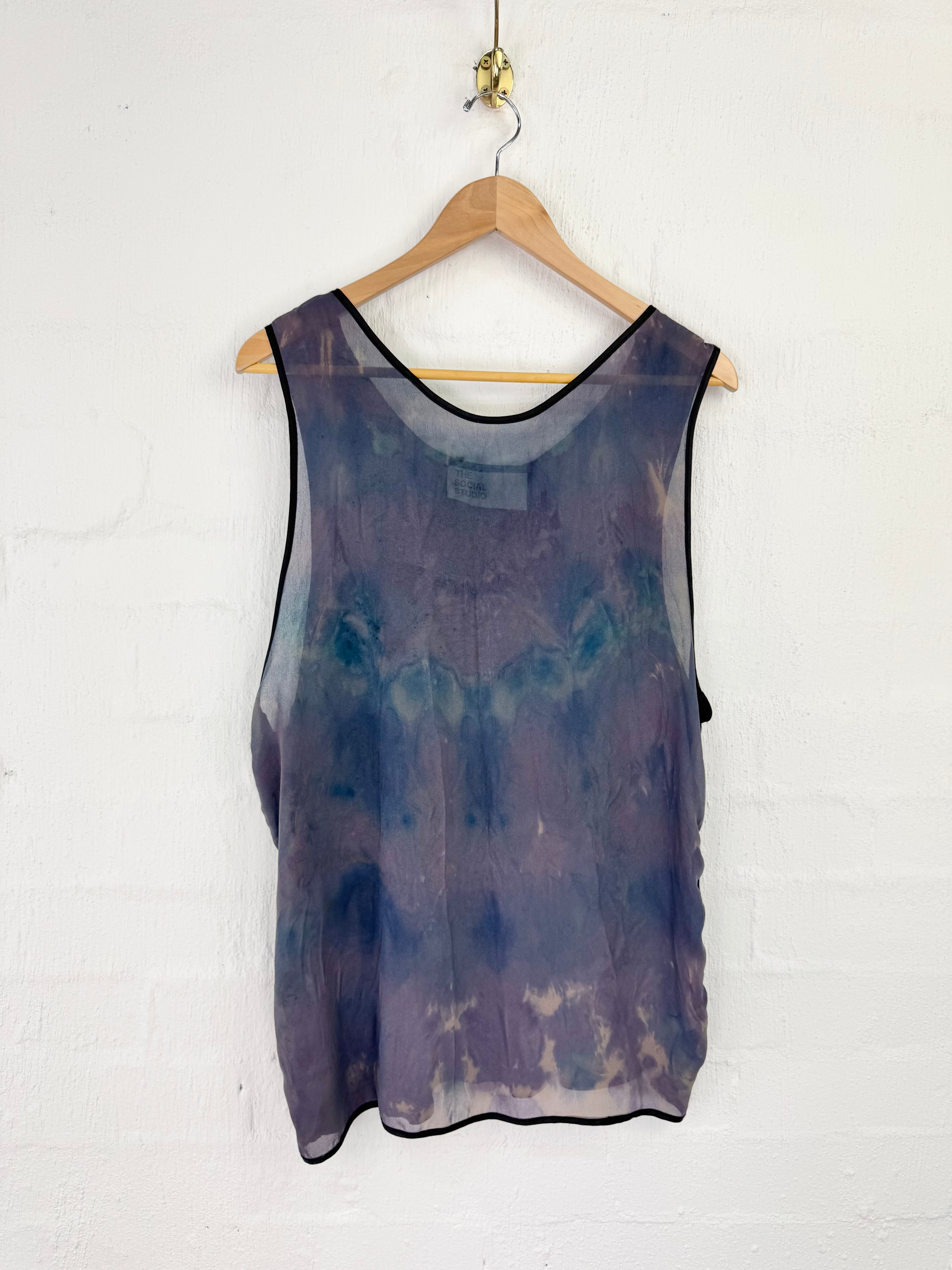 A sleeveless sheer mesh tank top with a purple and blue tie-dye pattern, hanging on a wooden hanger against a white painted brick wall.