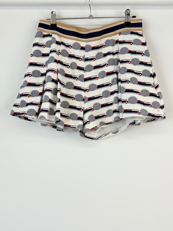 A pair of tennis-print shorts hanging against a white background, featuring a geometric pattern with tennis racket and ball motifs.