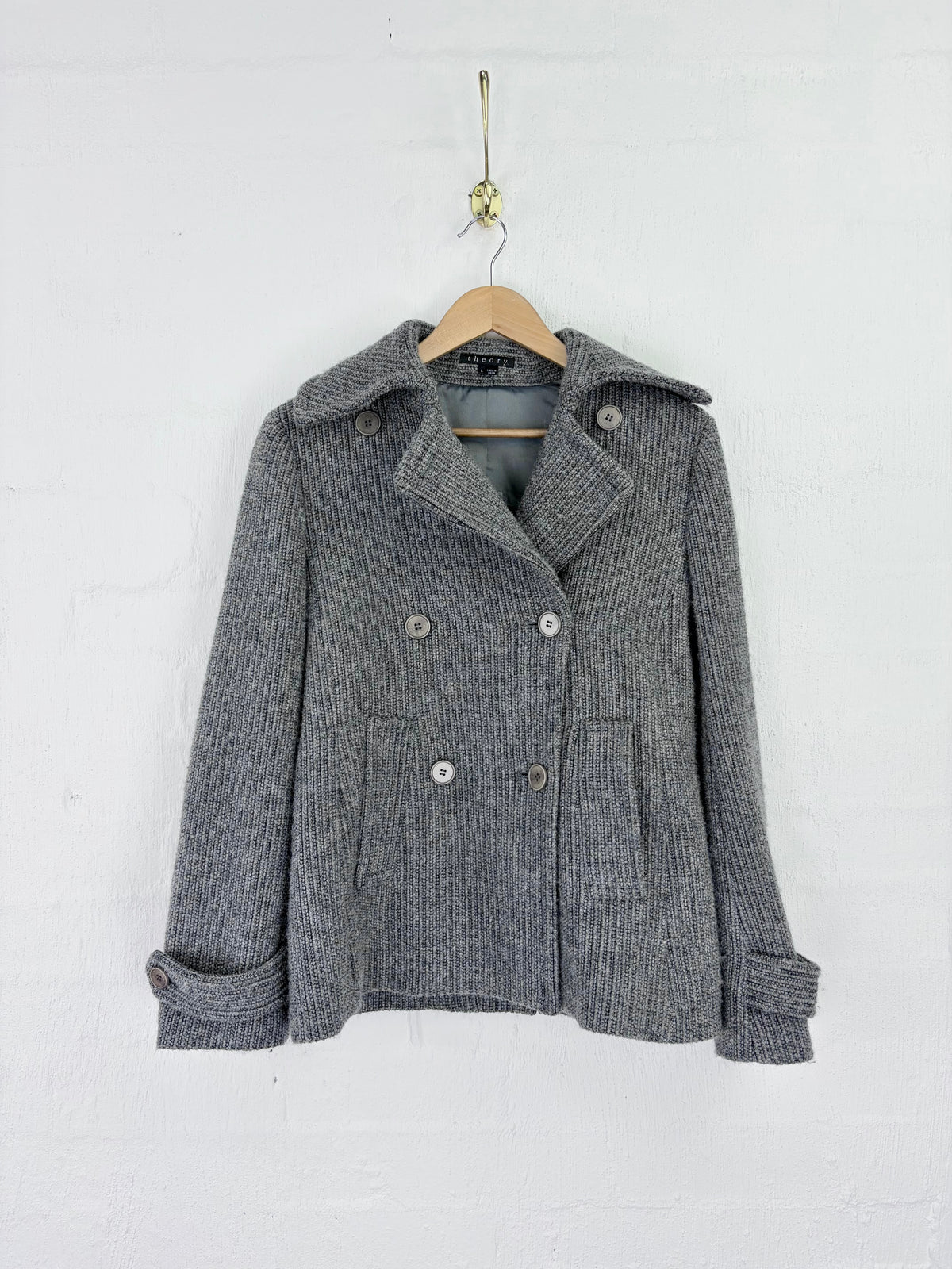 A gray ribbed double-breasted coat with a wide collar and silver buttons, hanging on a wooden hanger against a white painted brick wall.