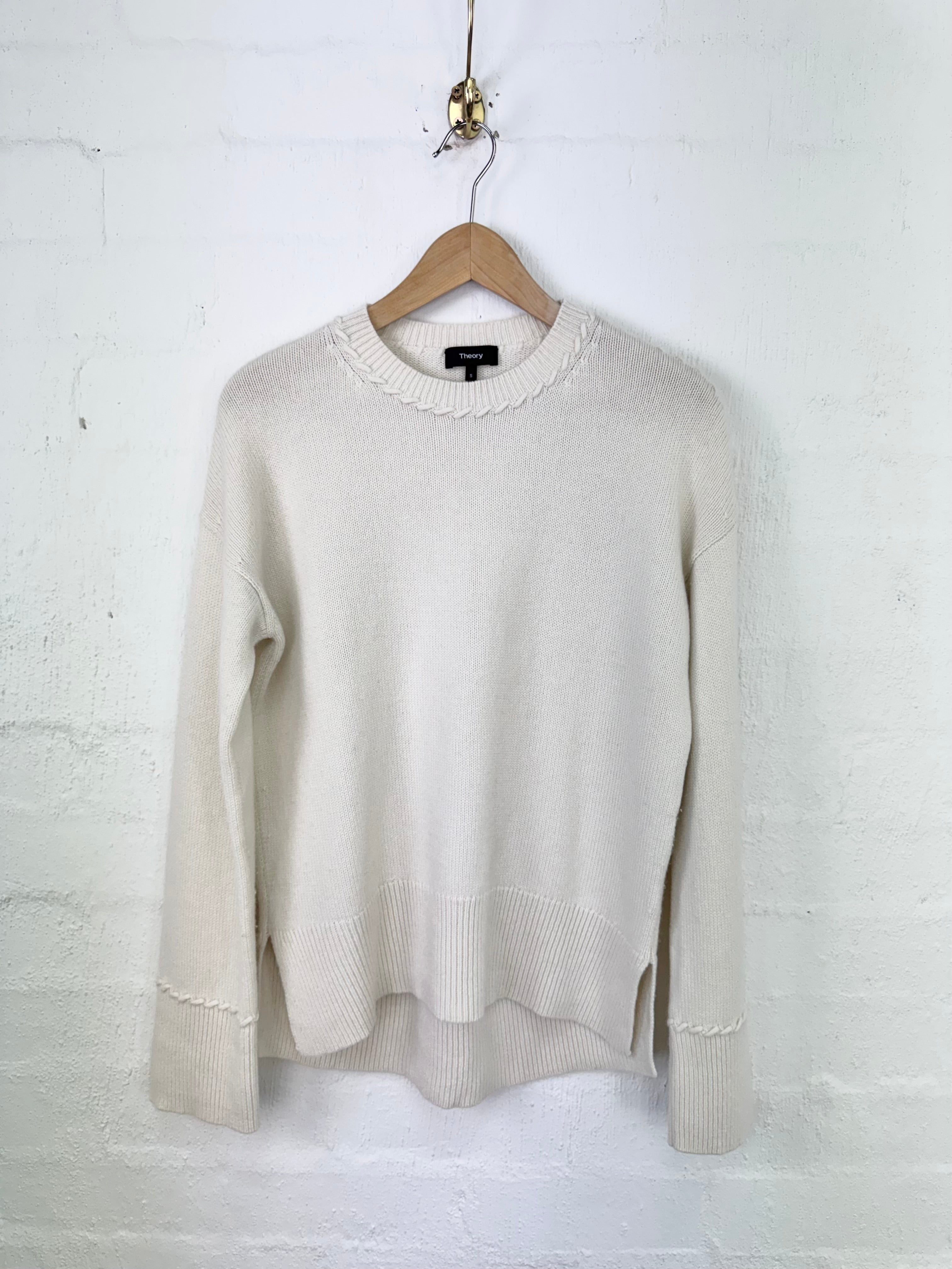 An off-white knit sweater with a scalloped crew neck and ribbed hem/cuffs, hanging on a wooden hanger against a white painted brick wall.