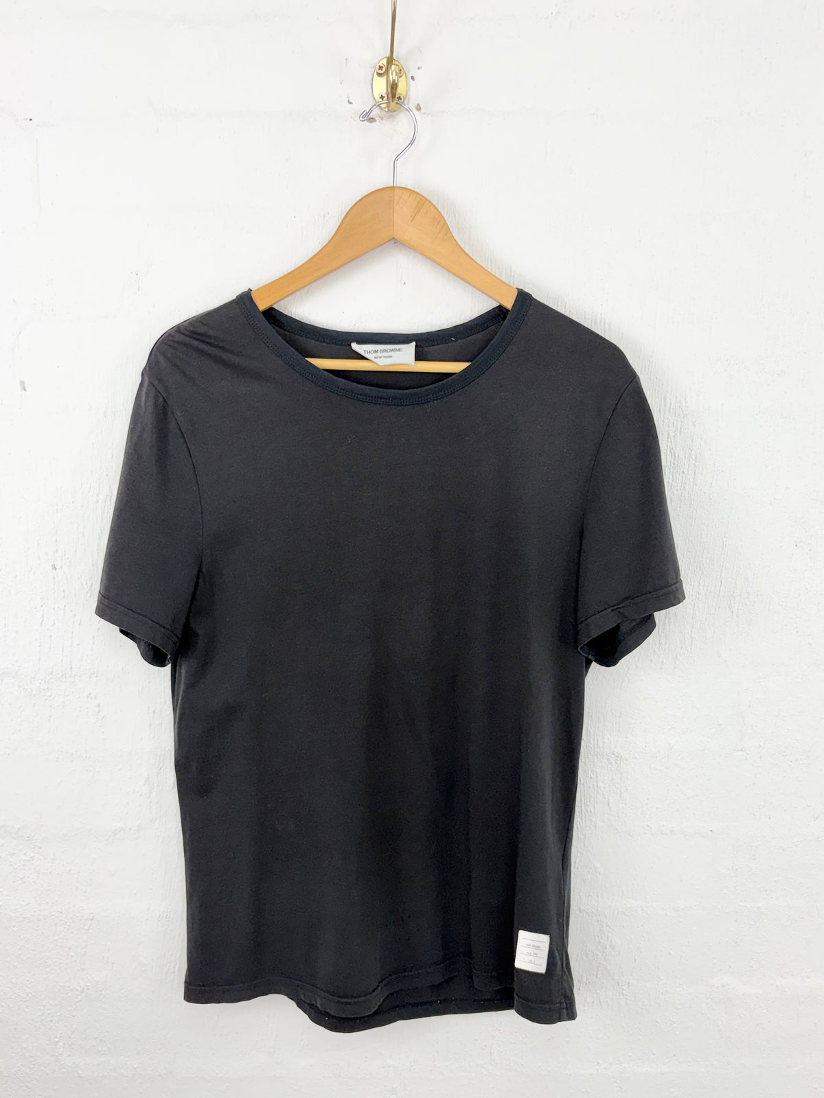A black short-sleeve crewneck t-shirt hanging on a wooden hanger against a white painted brick wall.