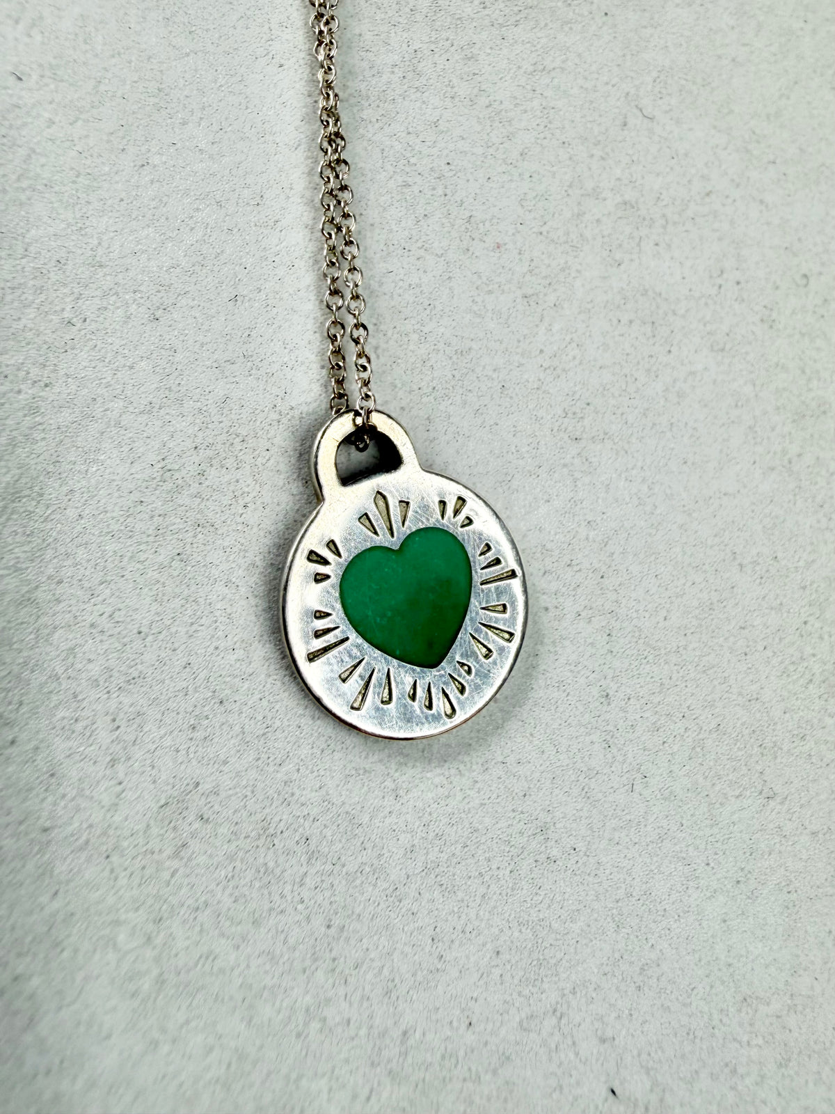A silver oval pendant necklace featuring a textured silver border with radiating cutouts and a vibrant green heart-shaped center, suspended from a silver chain against a light gray textured background.