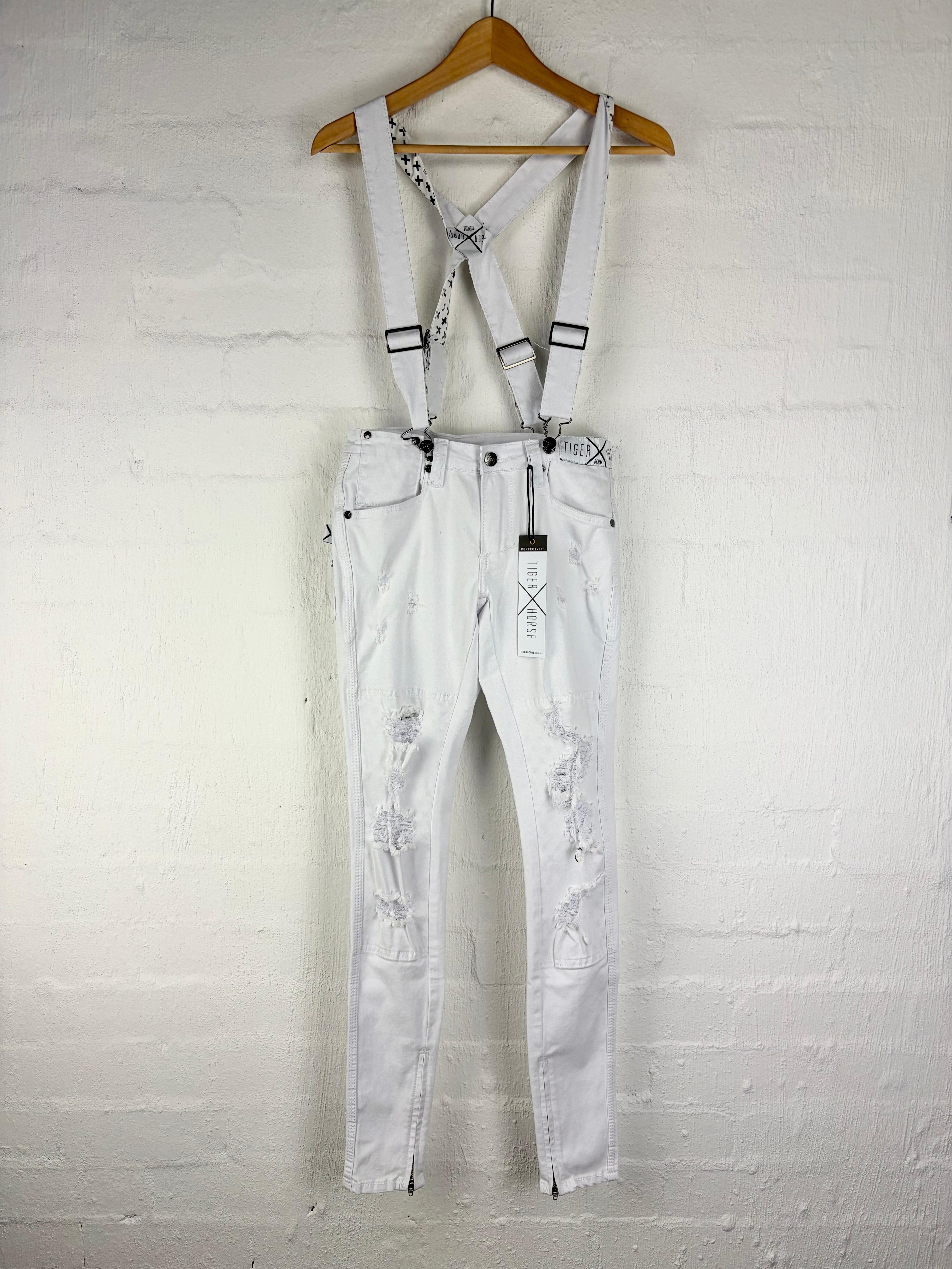 White distressed denim bib overalls with adjustable suspenders, hanging on a wooden hanger against a white painted brick wall.