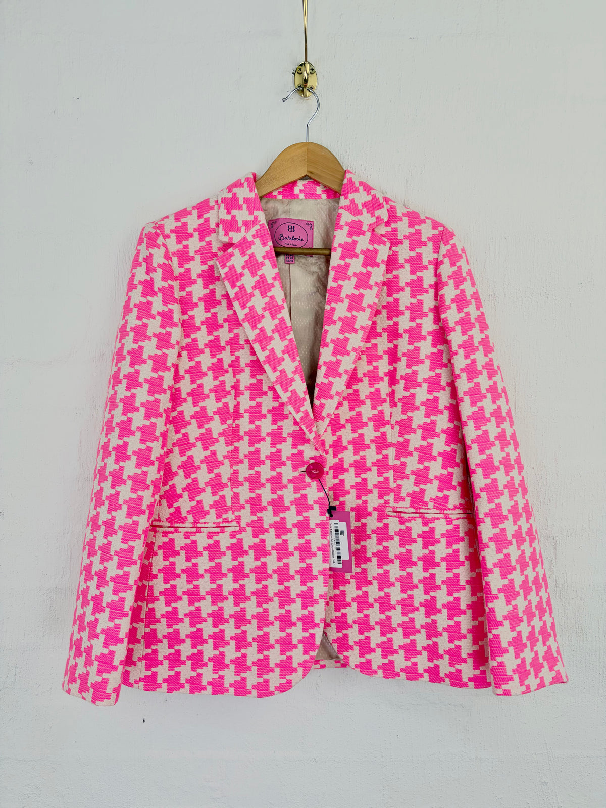 A pink and white houndstooth blazer jacket hanging on a wooden hanger against a white painted brick wall.