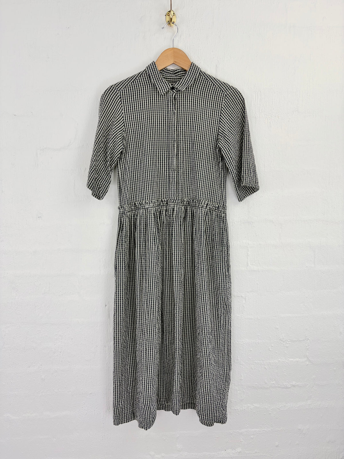 A short-sleeve collared dress with a black and white gingham pattern, hanging on a wooden hanger against a white brick wall.