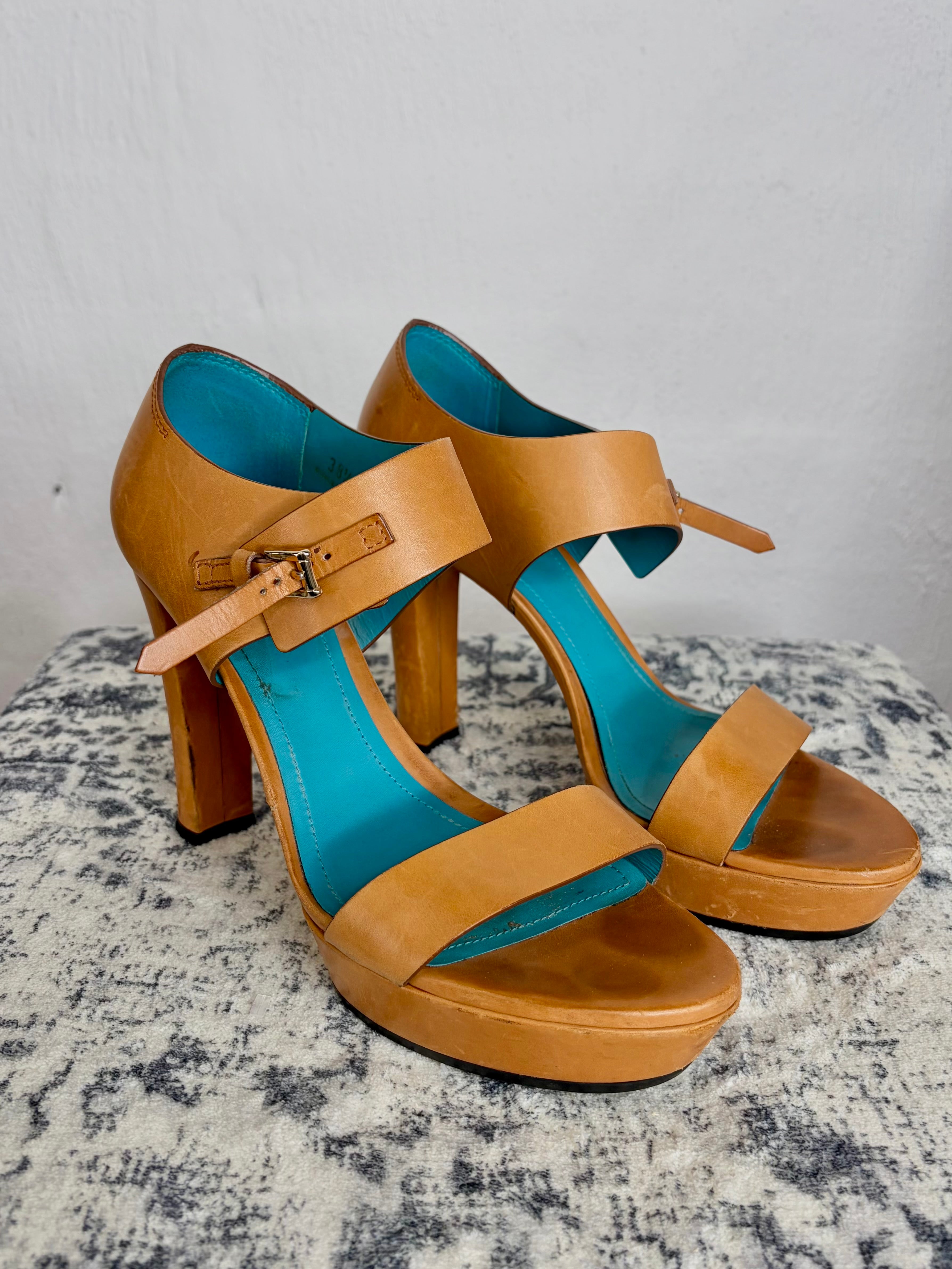 A pair of tan leather high-heeled sandals with turquoise blue insoles, displayed on a textured gray surface against a light gray wall.