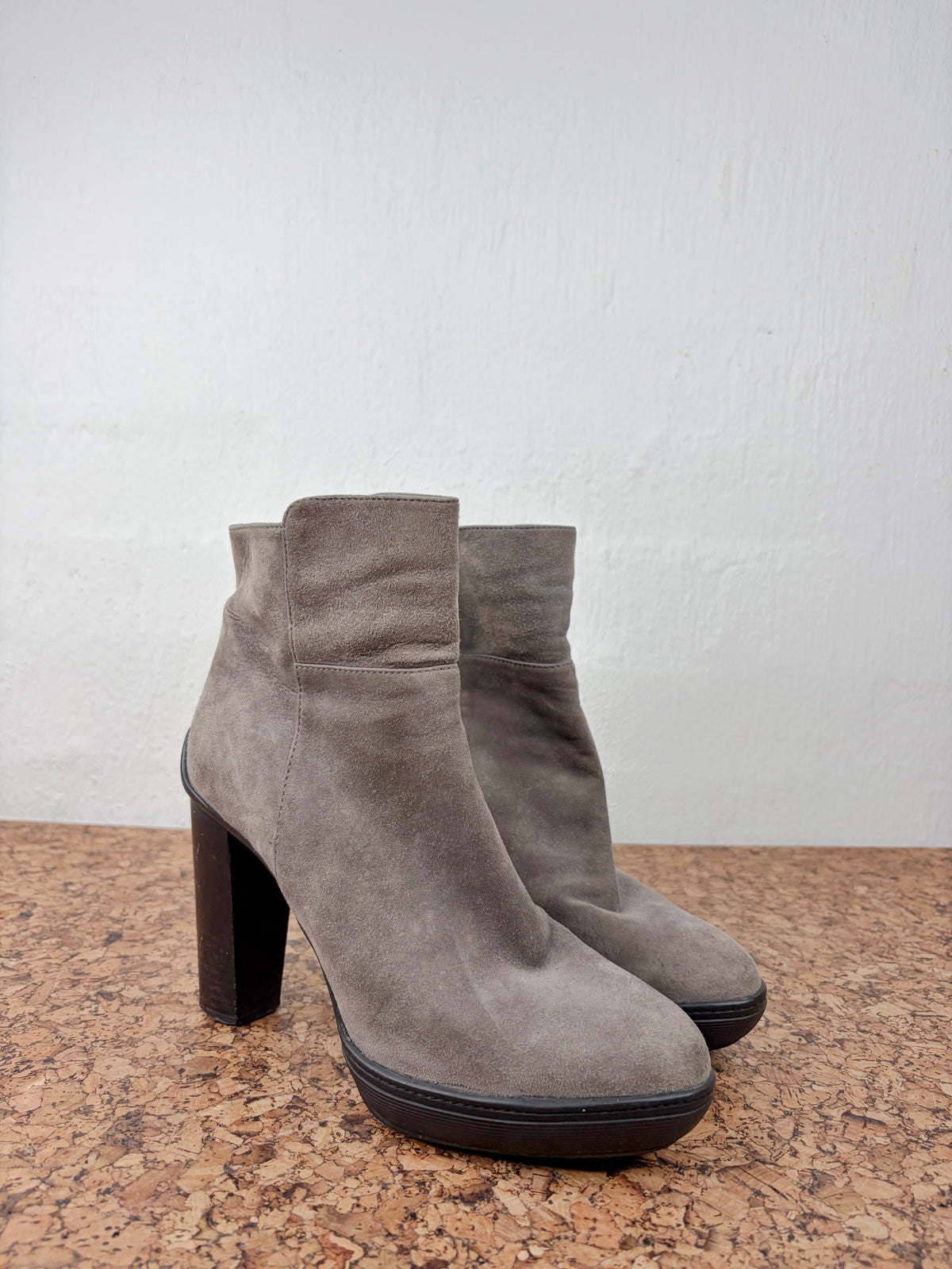 A pair of taupe-brown suede ankle boots with chunky block heels, displayed on a cork surface against a white wall.