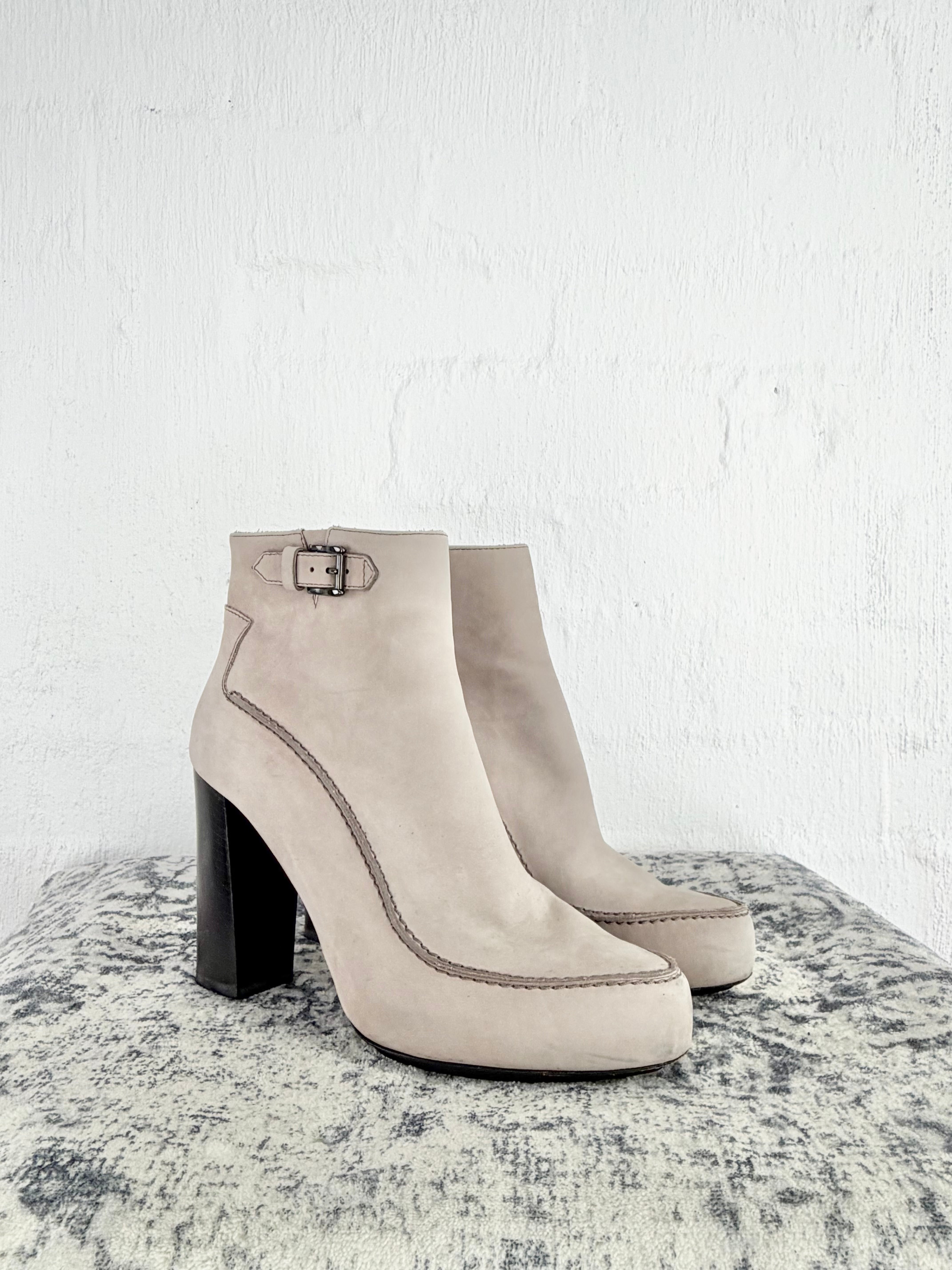 A pair of light beige suede ankle boots with chunky black block heels and a small buckle detail on the side, displayed on a textured gray surface against a white brick wall.