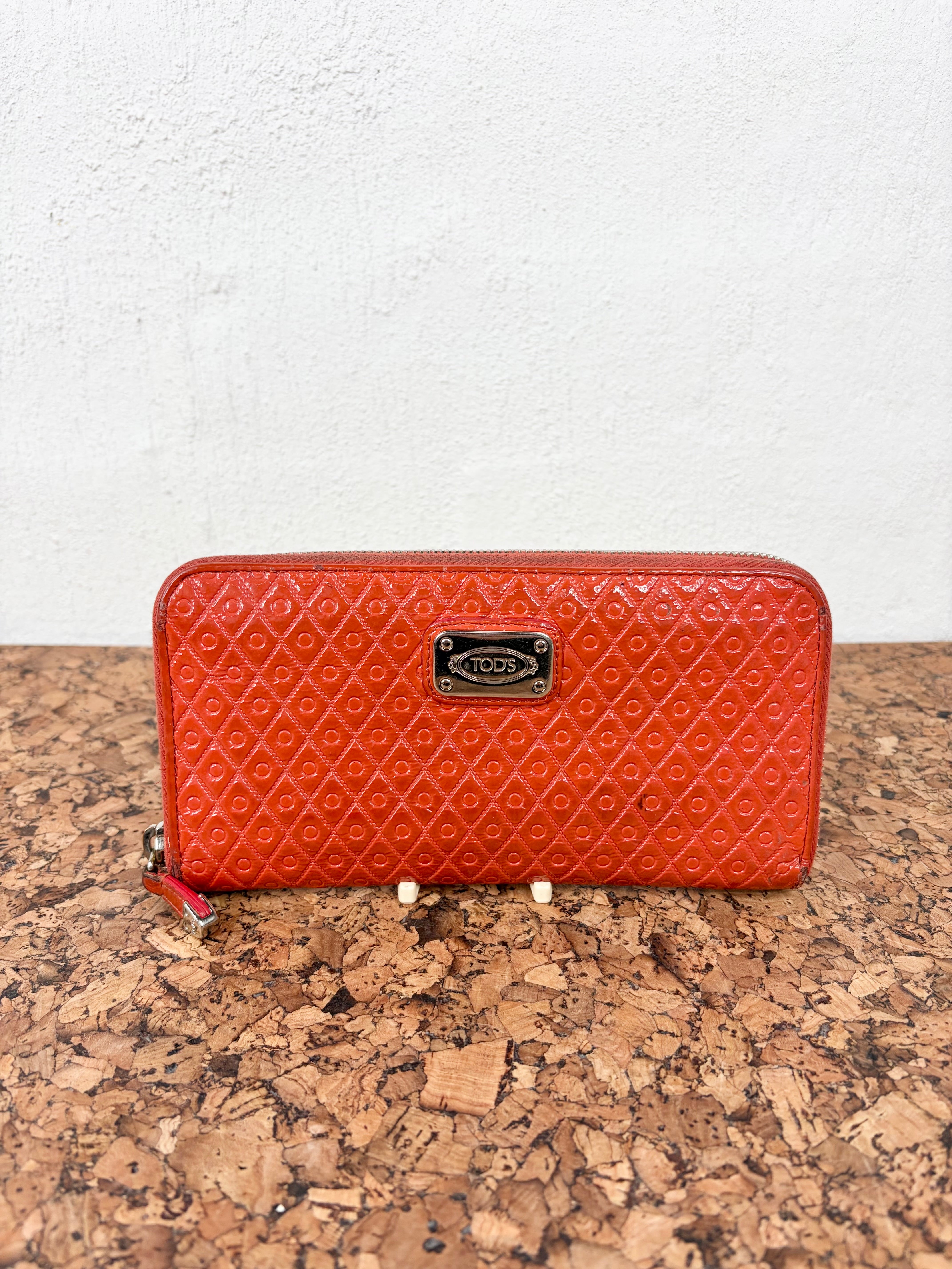 A bright orange-red embossed leather zip-around wallet with a silver-tone metal logo plate, resting on a cork surface against a white wall.