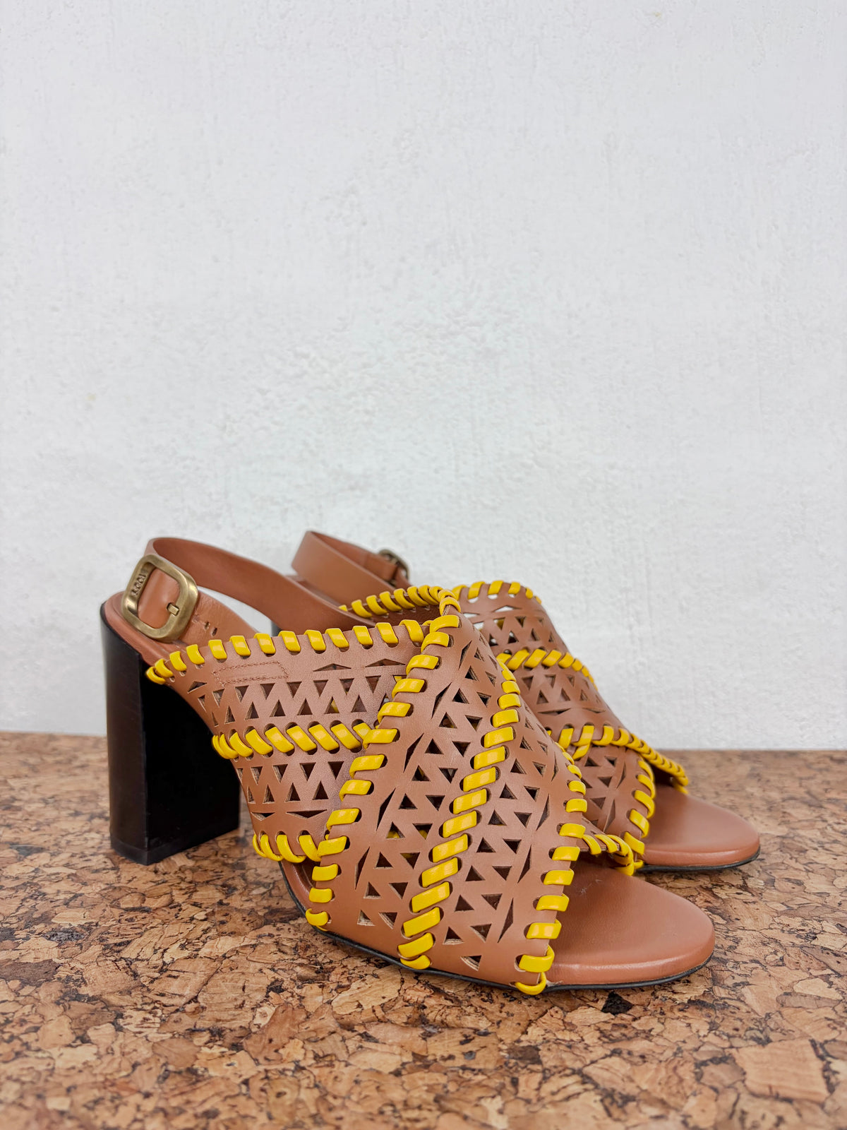 A pair of women's strappy heeled sandals with a tan leather base, geometric cutout pattern, and bright yellow braided accent details, shown on a cork surface against a white wall.
