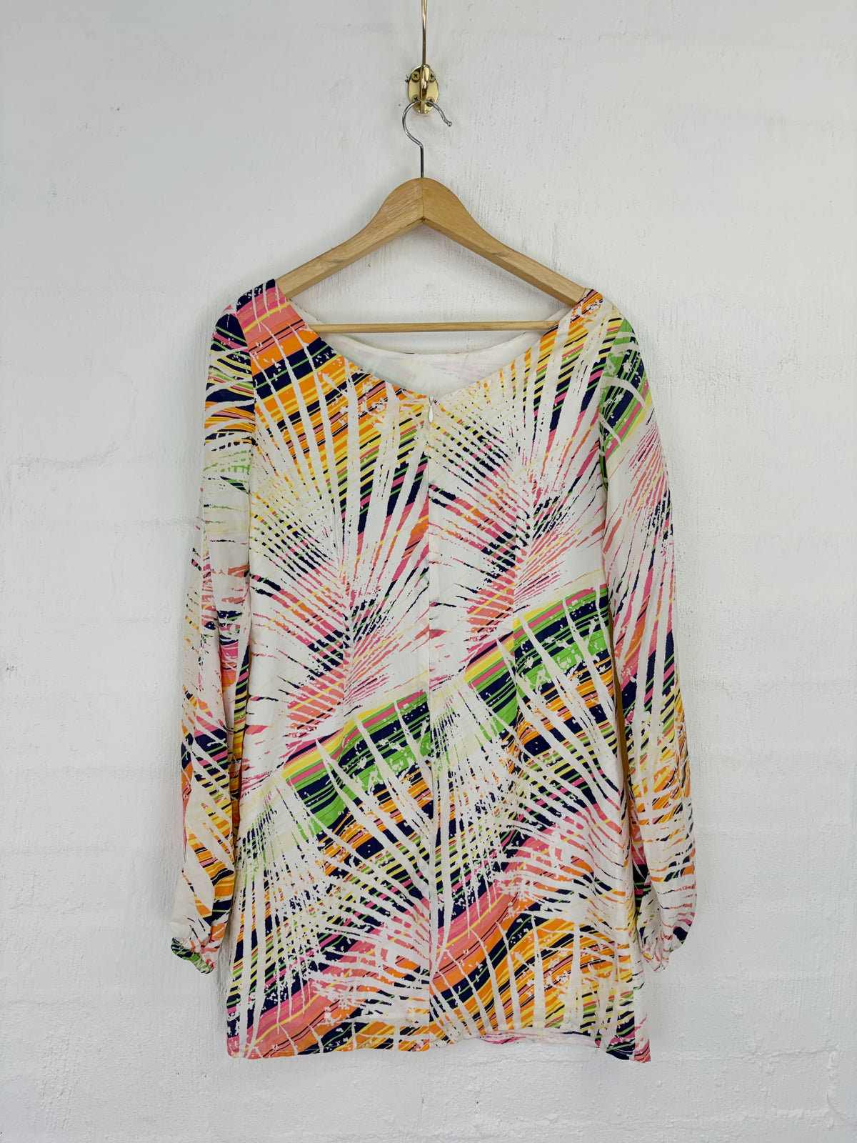A long-sleeve V-neck top with a vibrant multicolor abstract palm leaf print, hanging on a wooden hanger against a white painted brick wall.