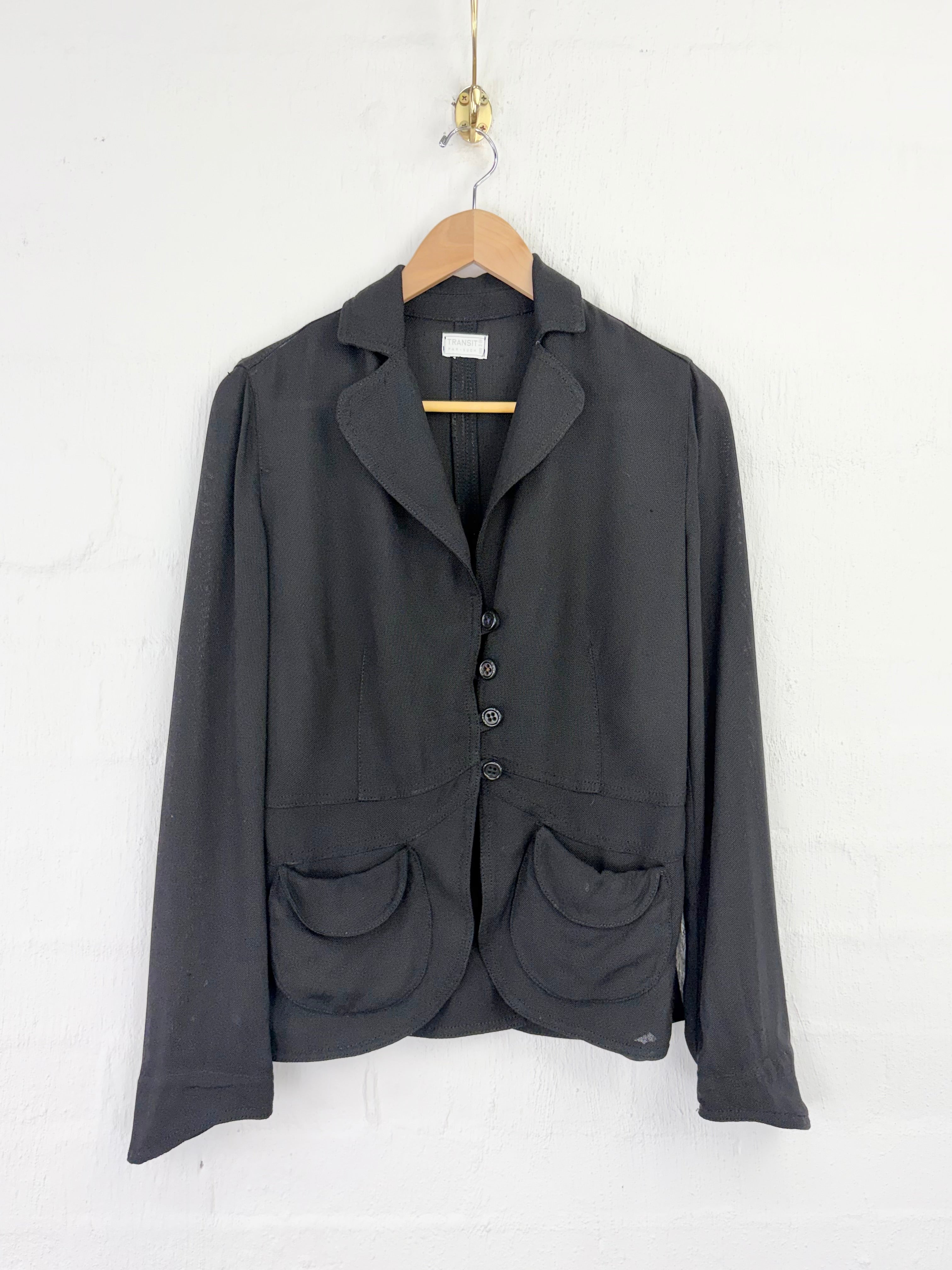 A dark charcoal gray blazer jacket with notch lapels, a three-button front closure, and two large front patch pockets, hanging on a wooden hanger against a white painted brick wall.