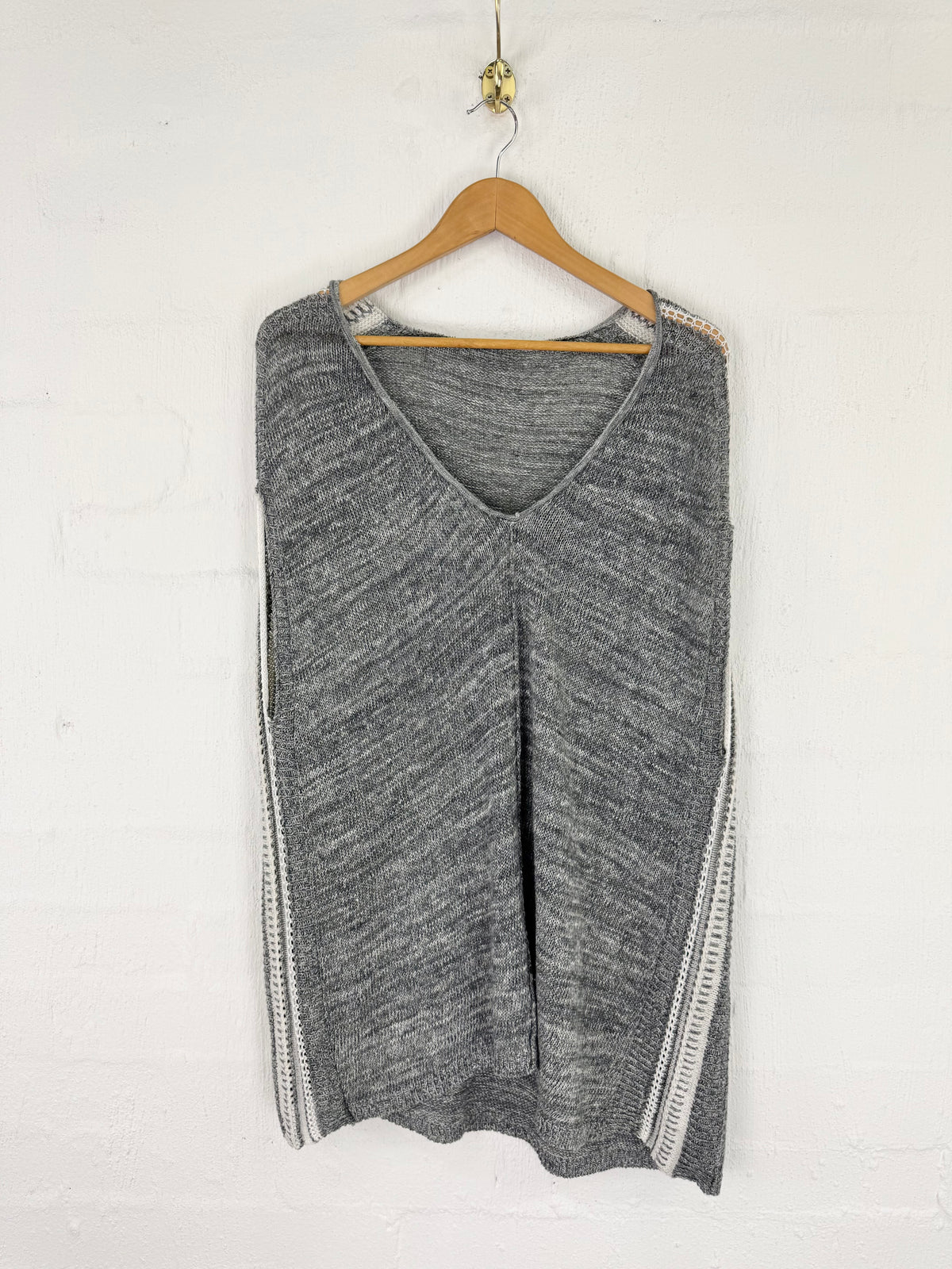 A gray knit sweater vest with a V-neck and textured side stripes, hanging on a wooden hanger against a white brick wall.