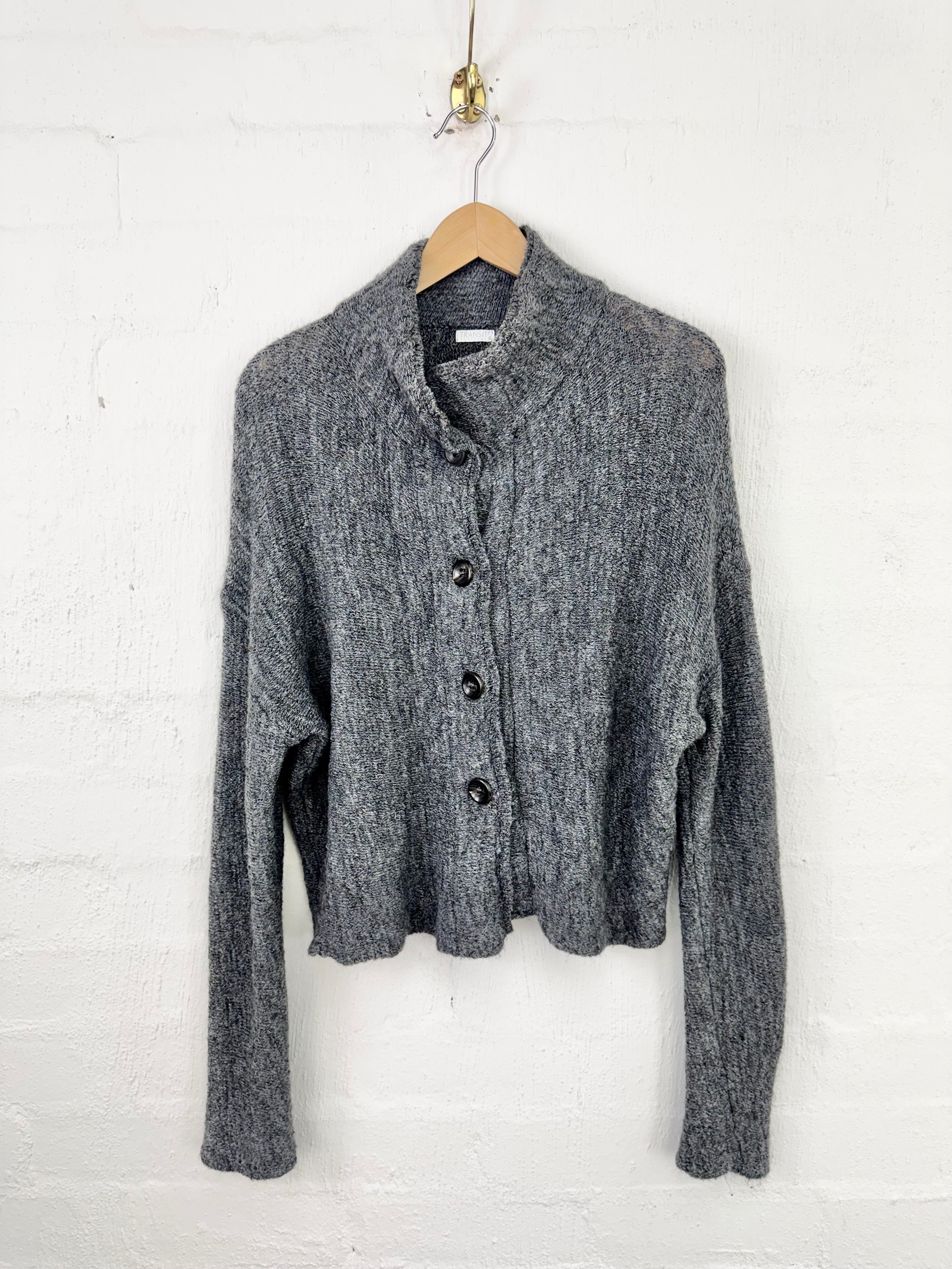 A gray knit cropped cardigan sweater with a high folded collar and front buttons, hanging on a wooden hanger against a white painted brick wall.