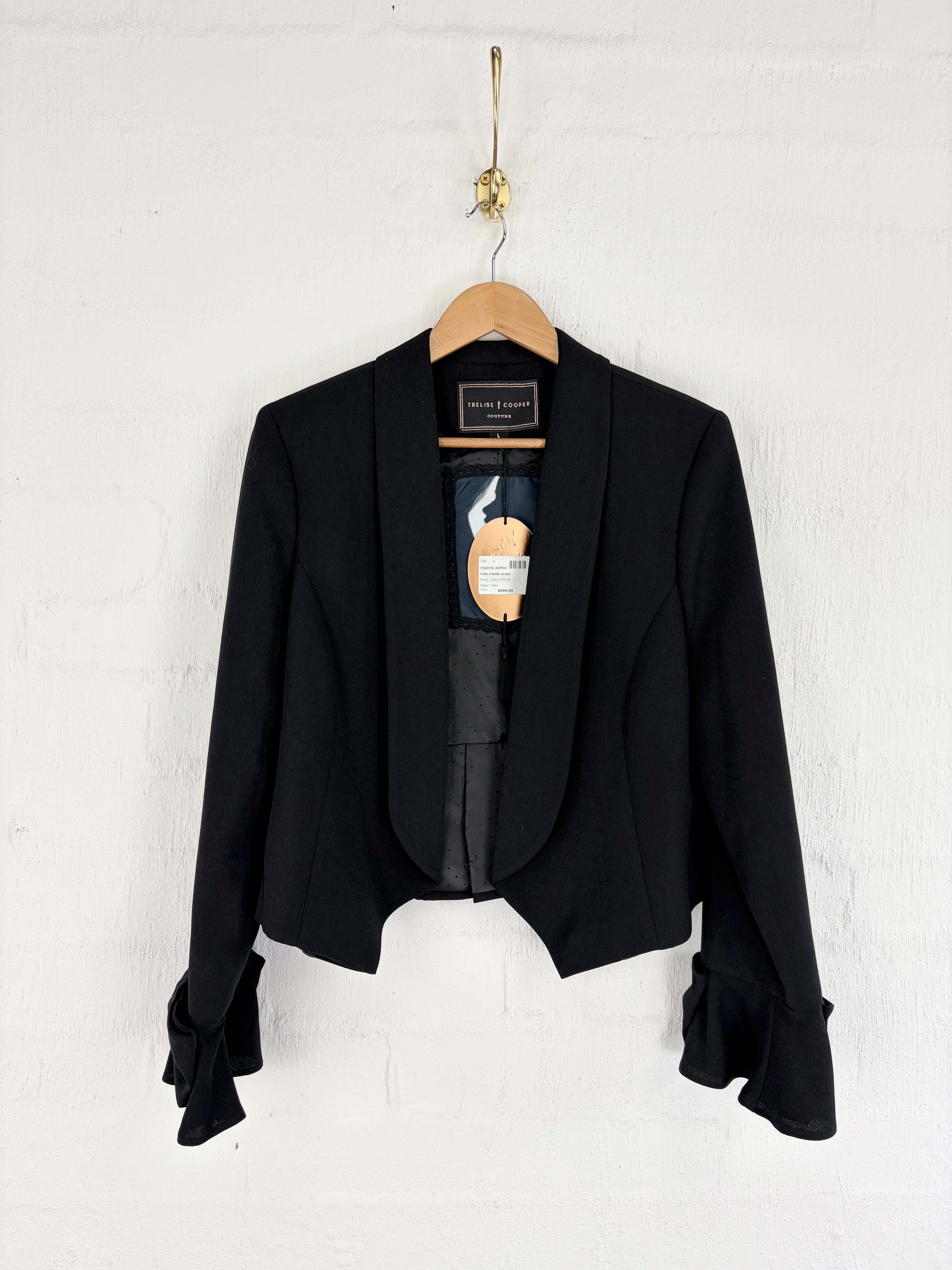 A black cropped blazer with dramatic ruffled sleeves, hanging on a wooden hanger against a white painted brick wall.