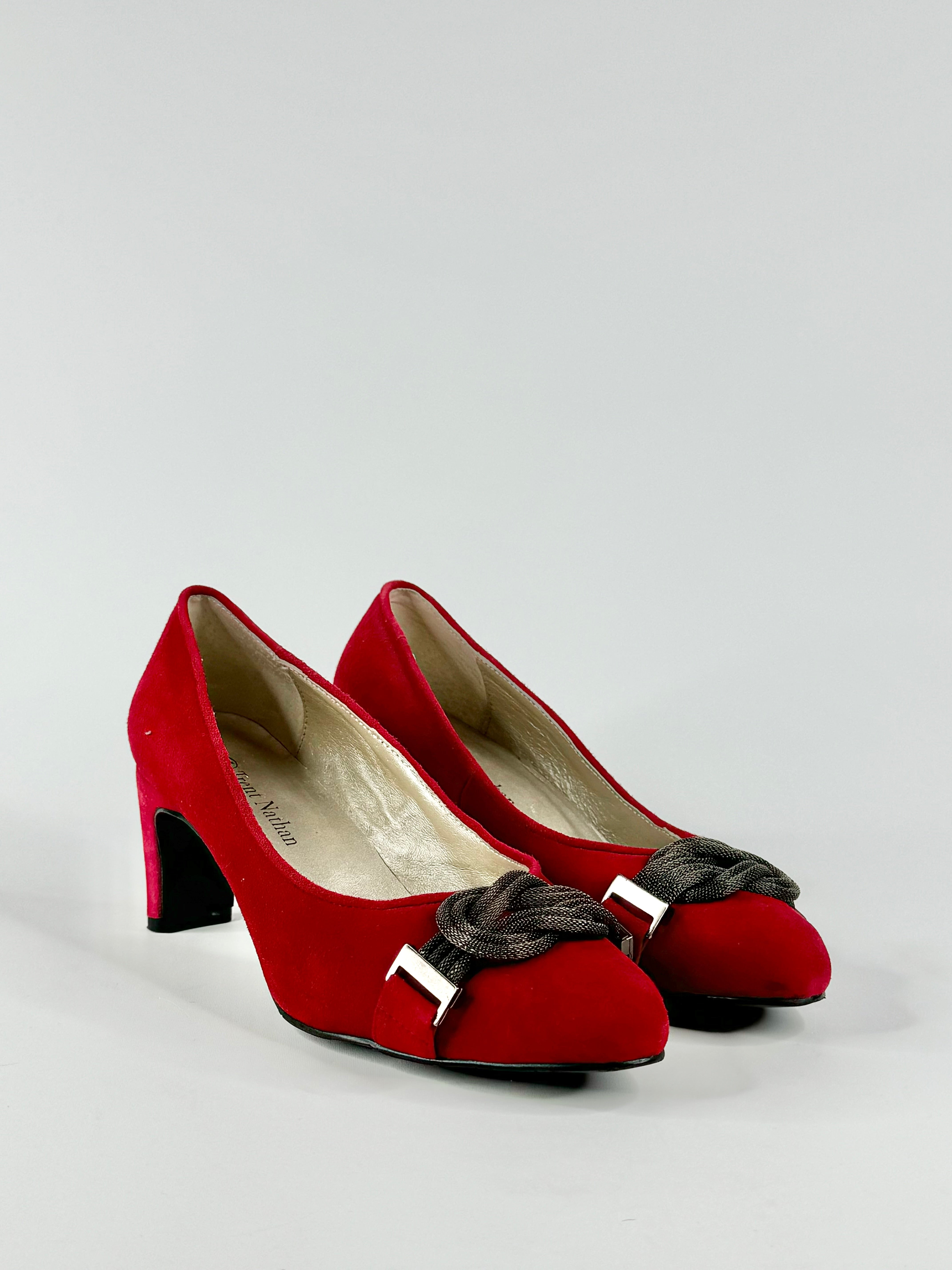 A pair of red suede heels with a small heel and a deep grey metal mesh knotted embellishment on the front.