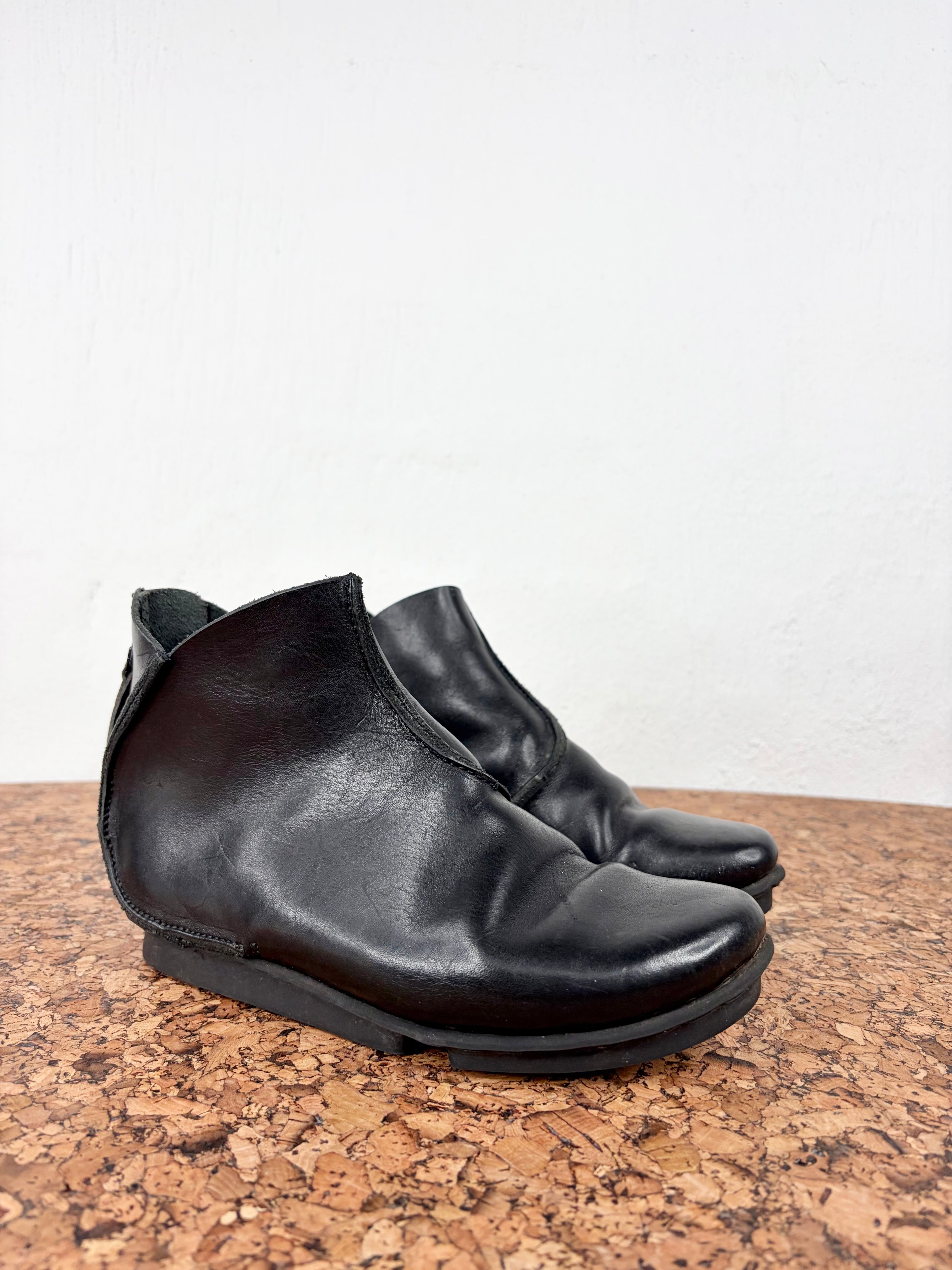 A pair of black leather ankle boots with a low block heel, resting on a cork surface against a white wall.