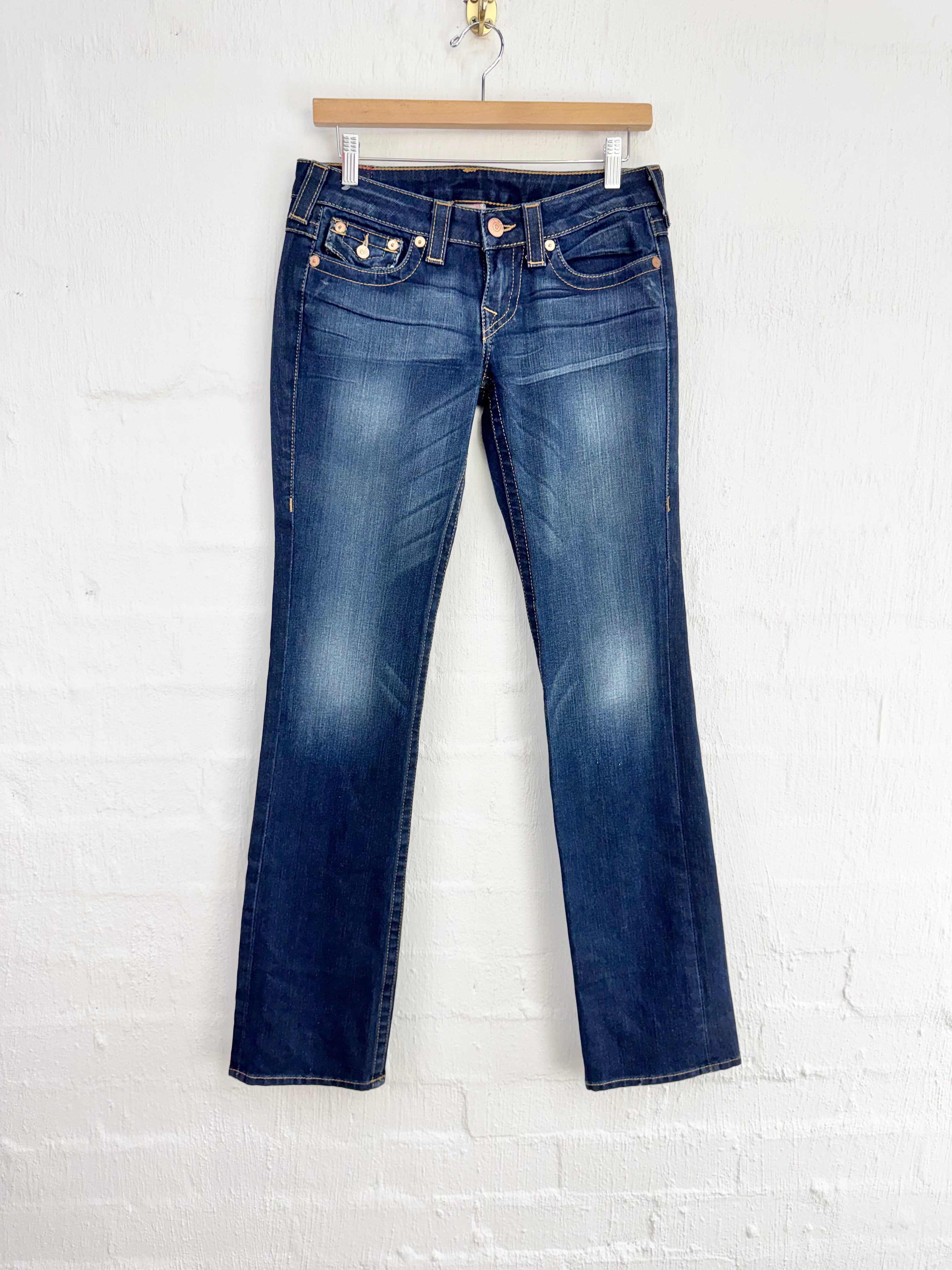 A pair of dark blue denim jeans with a slight flare, hanging from a wooden hanger against a white painted brick wall.
