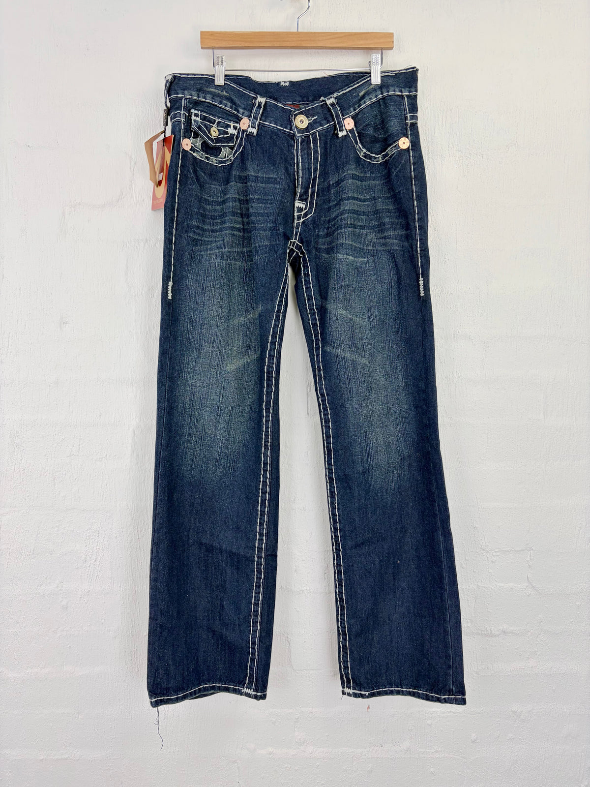 A pair of dark blue denim jeans with white contrast stitching, hanging on a wooden hanger against a white painted brick wall.