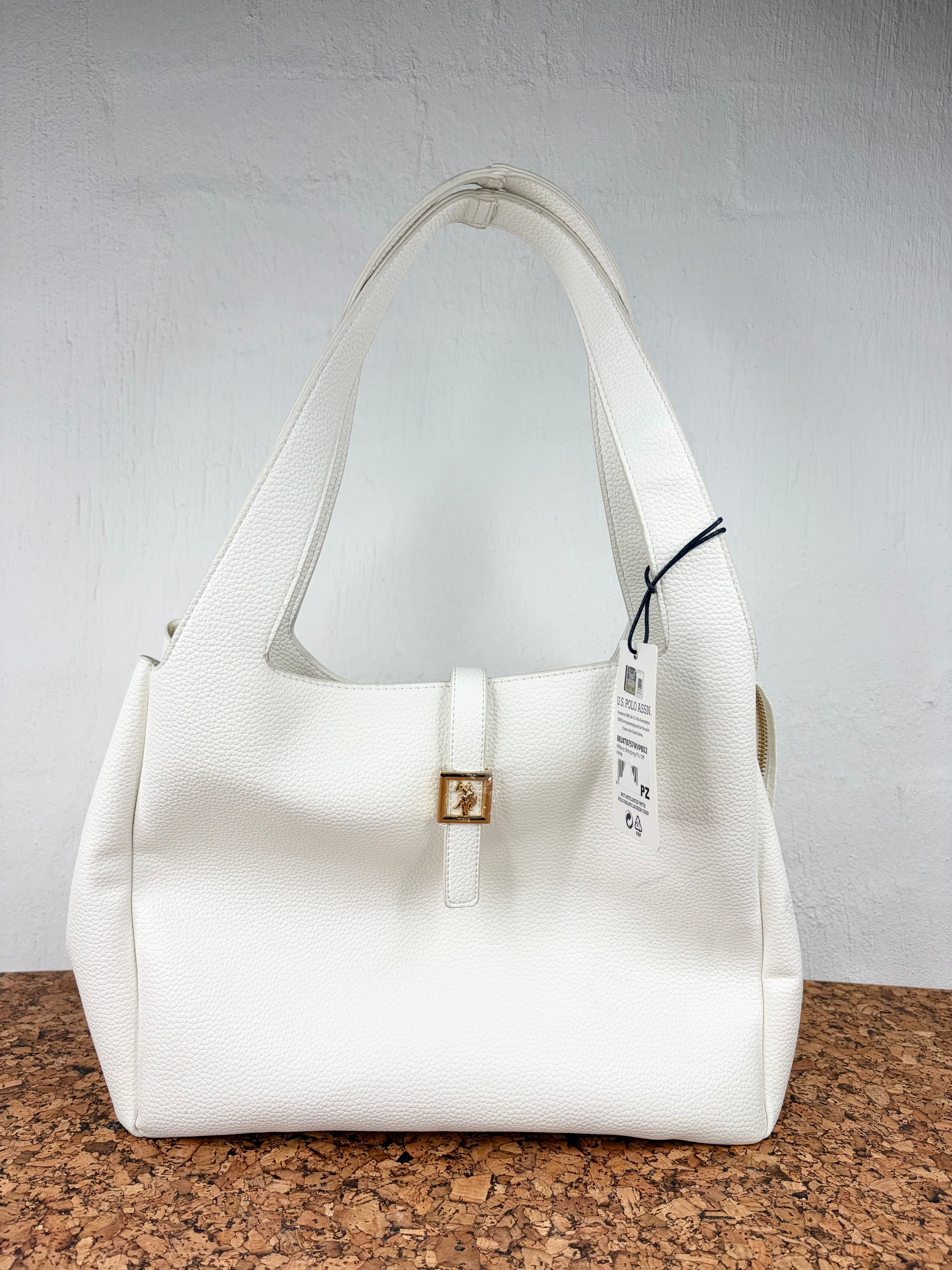 A white pebbled leather handbag with a small gold-tone logo clasp, resting on a cork surface against a white brick wall.