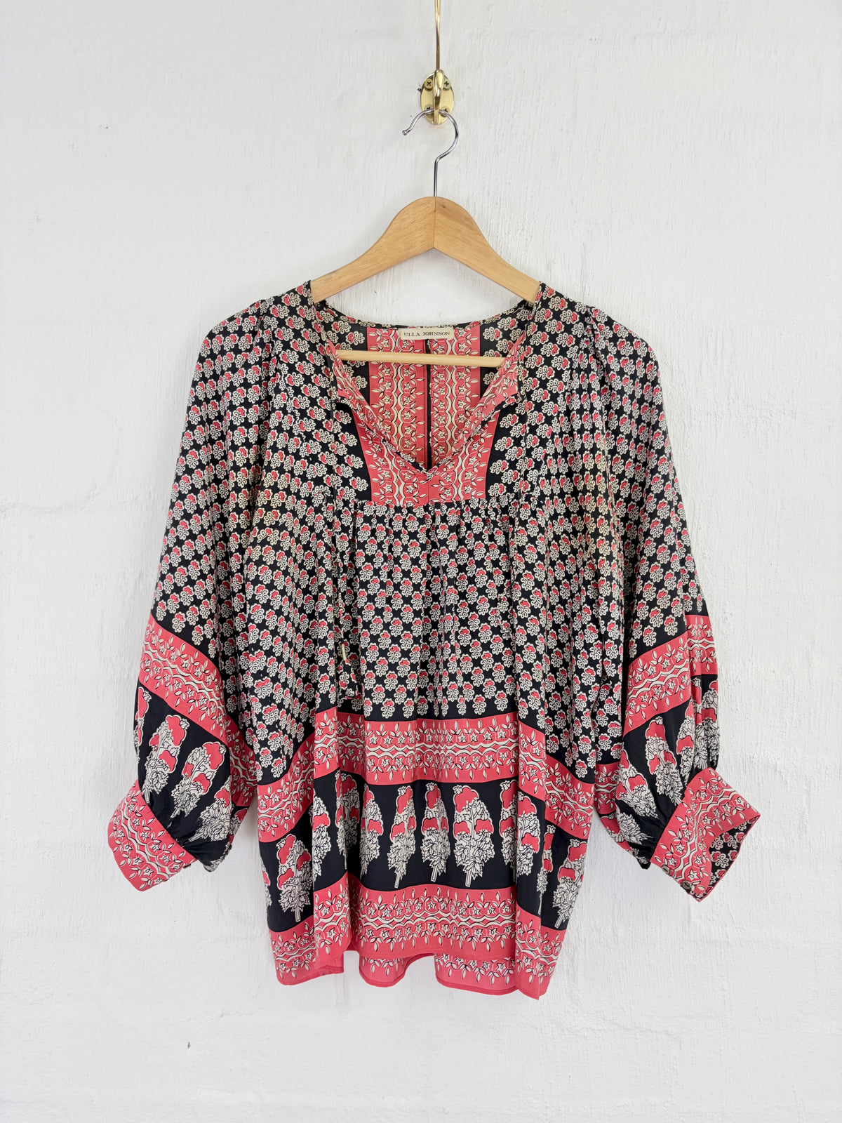 A bohemian-style long-sleeve top with a black, pink, and white geometric pattern, hanging on a wooden hanger against a white textured wall.